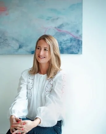 A smiling woman with shoulder-length blonde hair wearing a white blouse with lace details, sitting in front of an abstract blue and pink painting.