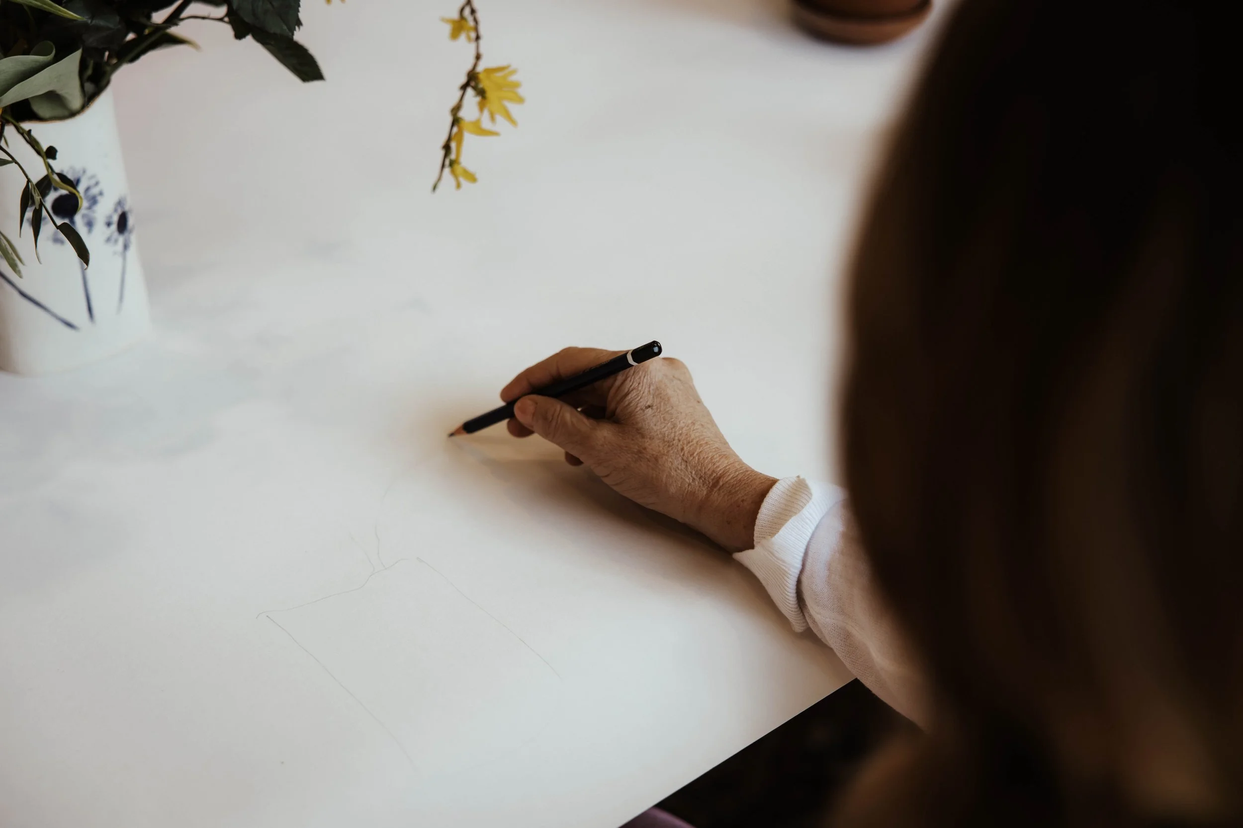 An elderly person drawing on a blank piece of paper with a black pencil at a white table, with plants in white pots nearby.