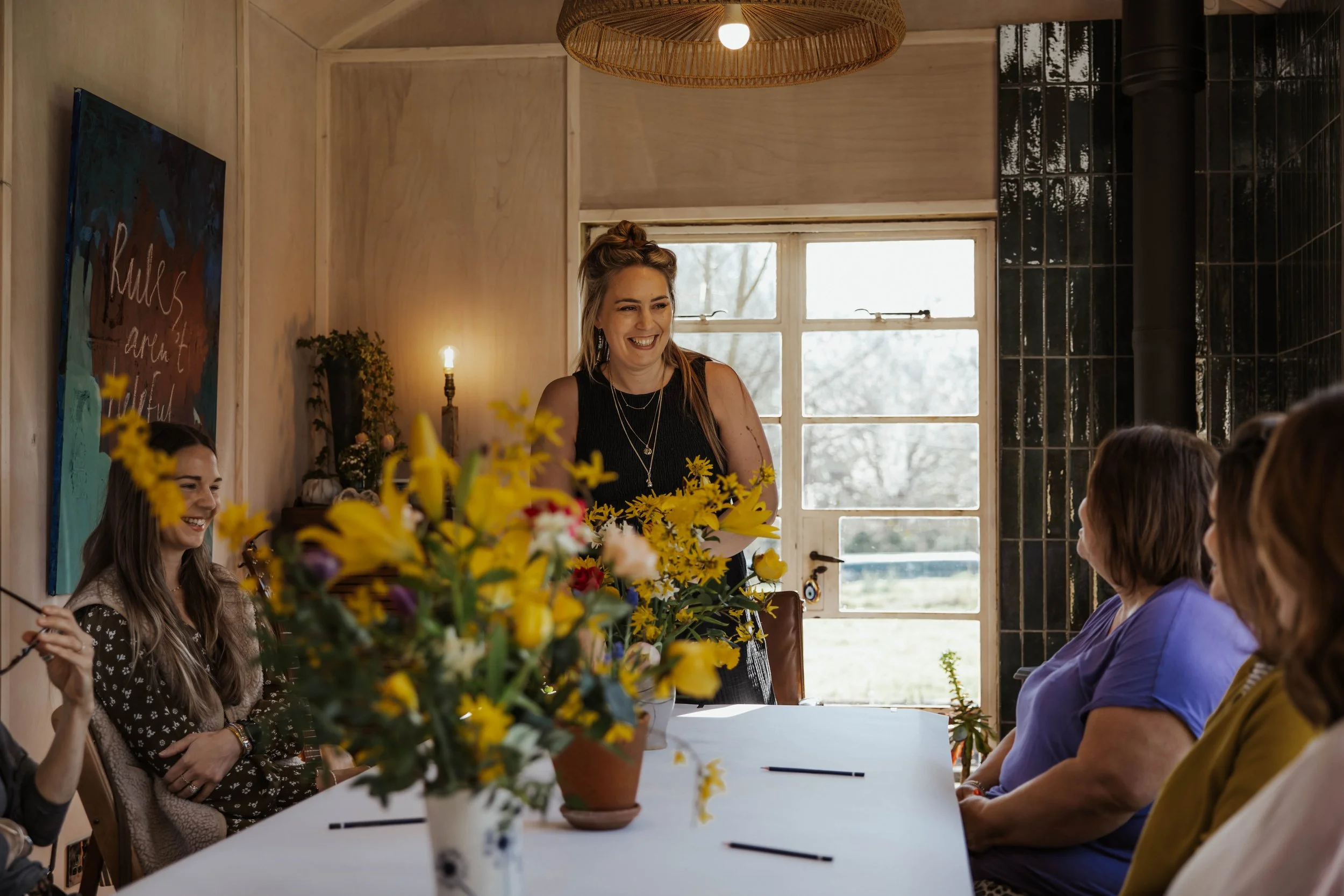 Women gathered around a table with a large bouquet of yellow flowers, an example of a communal painting session at A Creative Pause