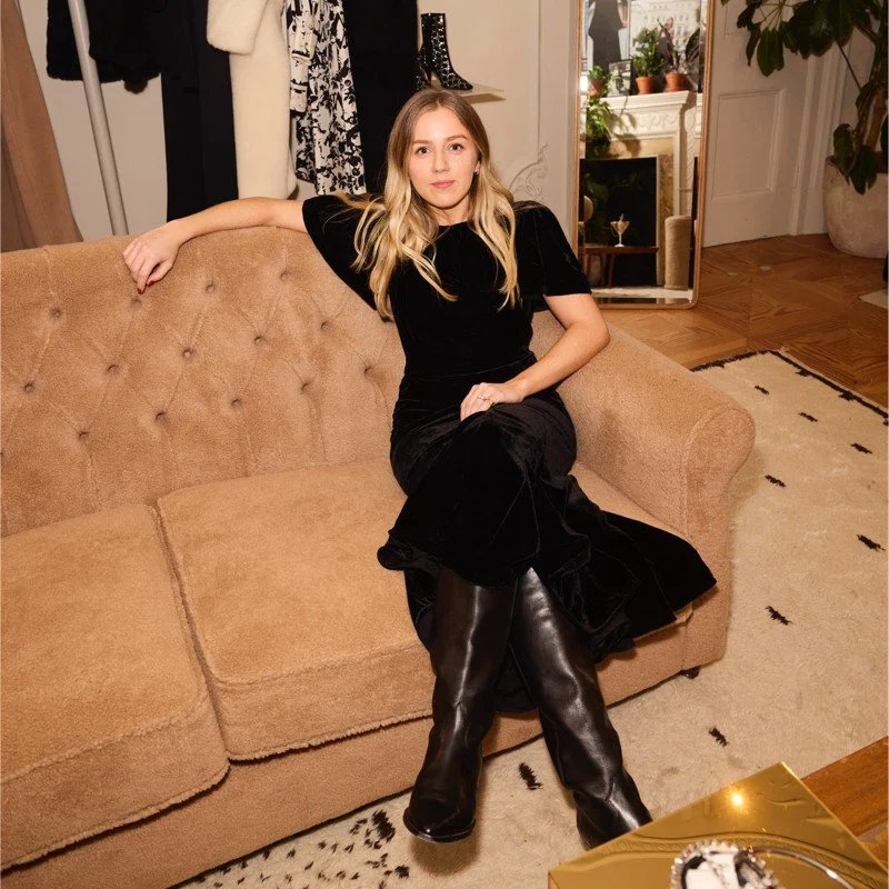 A young woman with blonde hair sitting on a beige sofa in a living room. She is wearing a black dress and tall black boots. There is a mirror behind her, reflecting some plants and furniture, and a white door open to another room. The room has a cream-colored carpet and wooden floor, with some plants and decorative items visible.