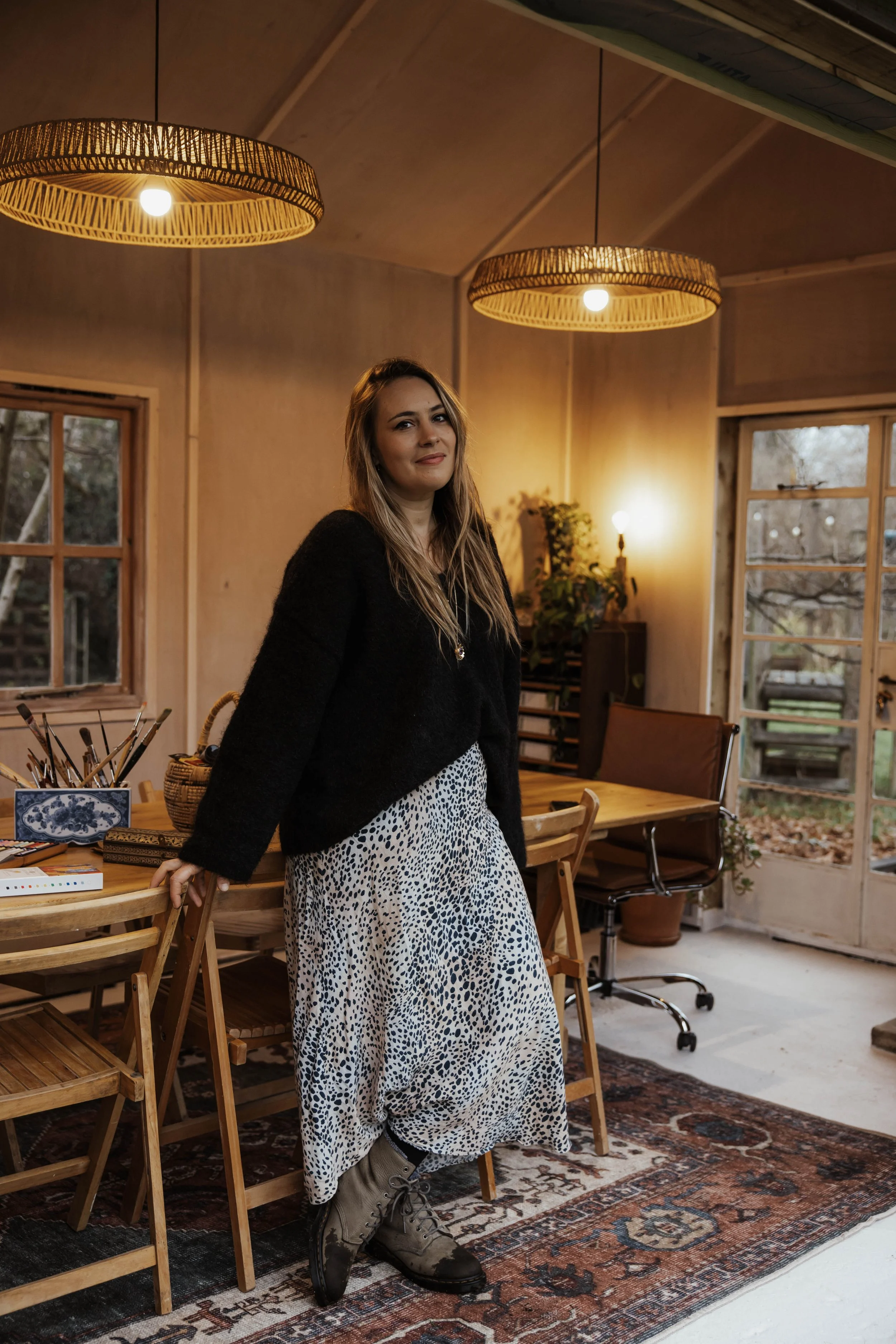 A woman standing in a cozy, well-lit room with wooden walls, next to a wooden dining table, wearing a black top and a white and black patterned skirt, with outdoor scenery visible through the windows and glass door.