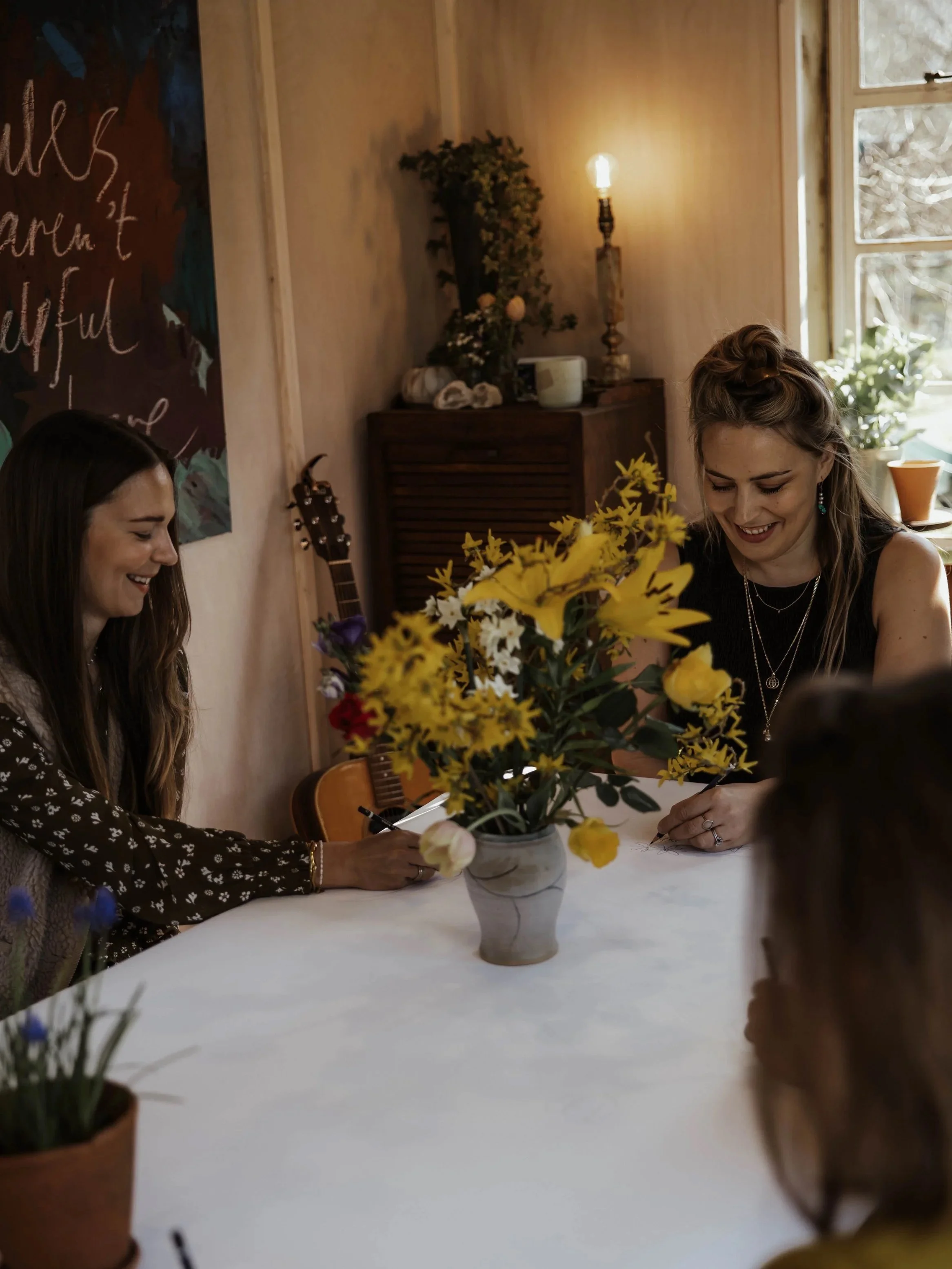 Women gathered around a table with a large bouquet of yellow flowers, an example of a communal painting session at A Creative Pause