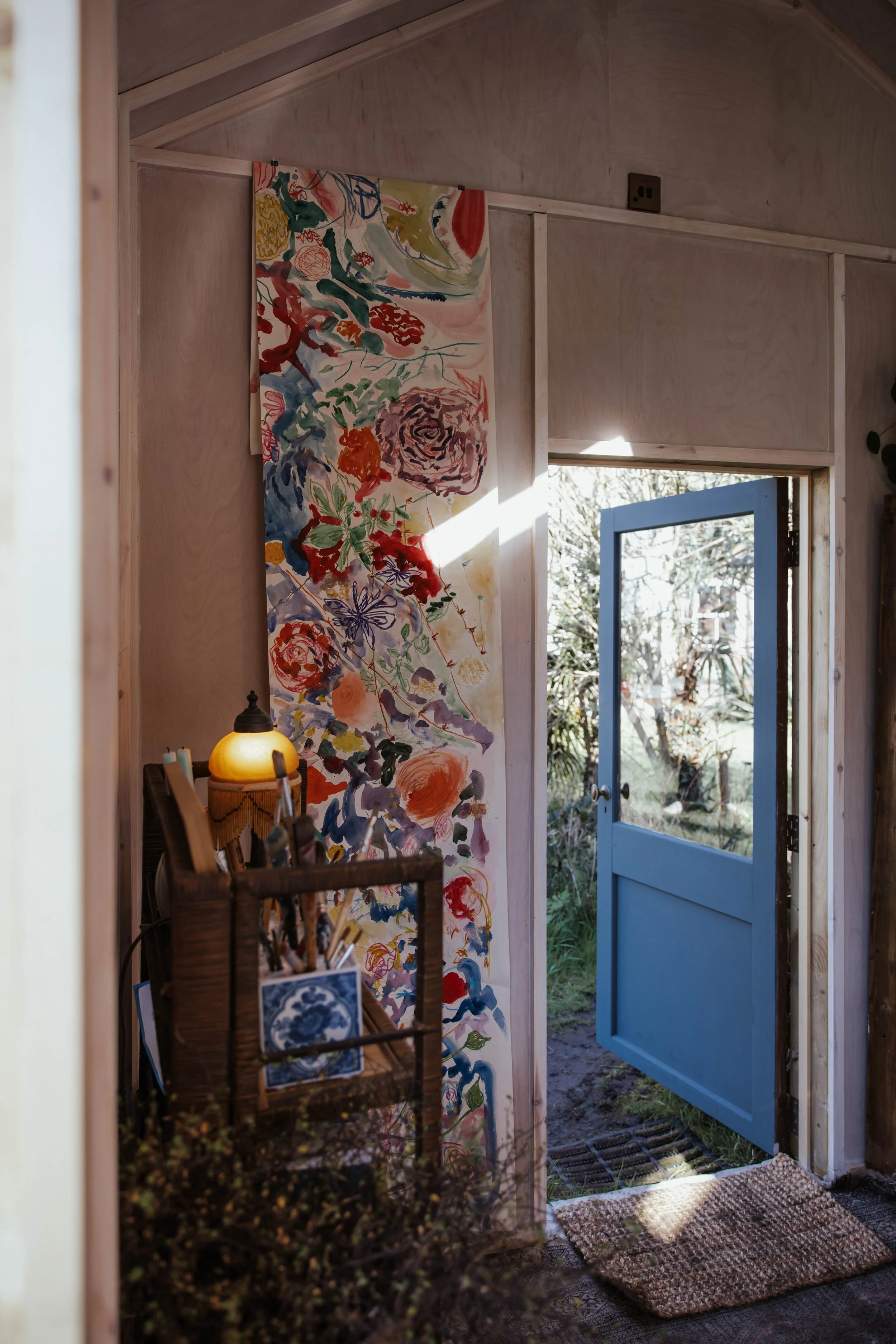 Interior view of a cozy room with a blue door open to a garden, a colorful floral painting on the wall, a small wooden shelf with art supplies, and a lamp, bathed in natural light. A Creative Pause art studio