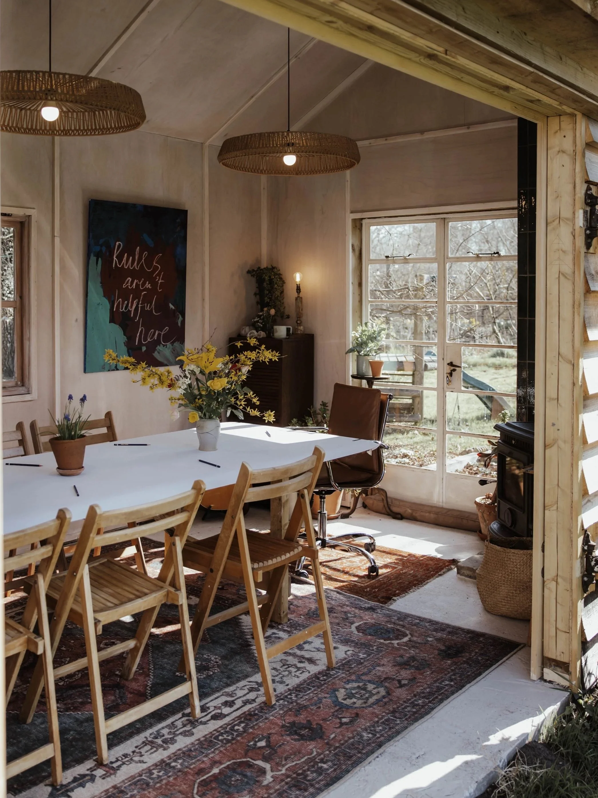 Cozy dining room with a wooden table, surrounded by wooden chairs and a leather office chair. A large window lets in natural light, overlooking an outdoor scene with trees. Decor includes potted plants, a vase with flowers, a vintage-style rug, and a black wood stove. Woven pendant lights hang from the ceiling, and a wall art piece reads 'Rules aren't helpful here'.