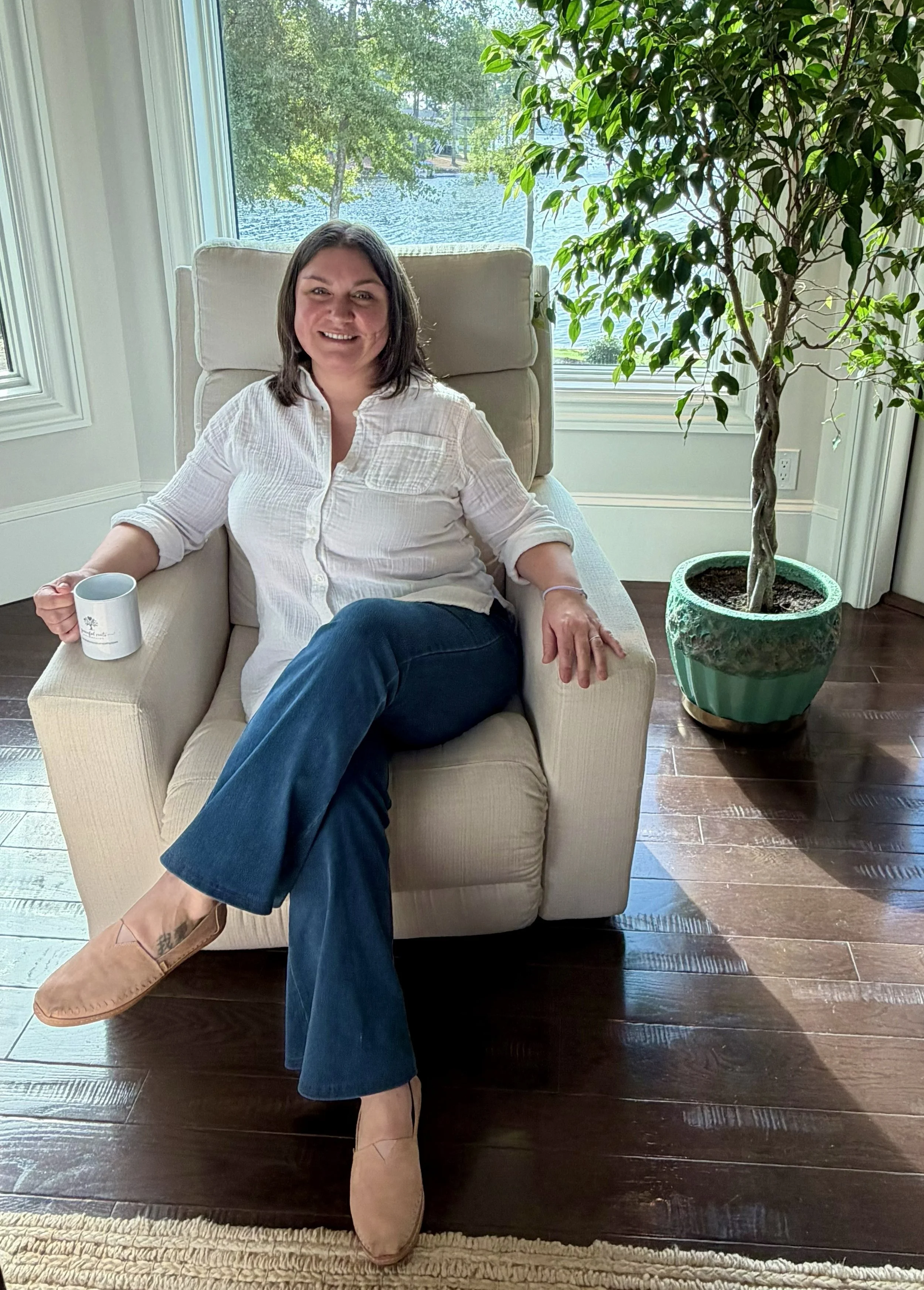 A woman sitting on a beige armchair in front of a large window with a view of a lake and trees, holding a white mug, wearing a white button-up shirt, dark blue jeans, and tan shoes, with a green potted plant beside her.