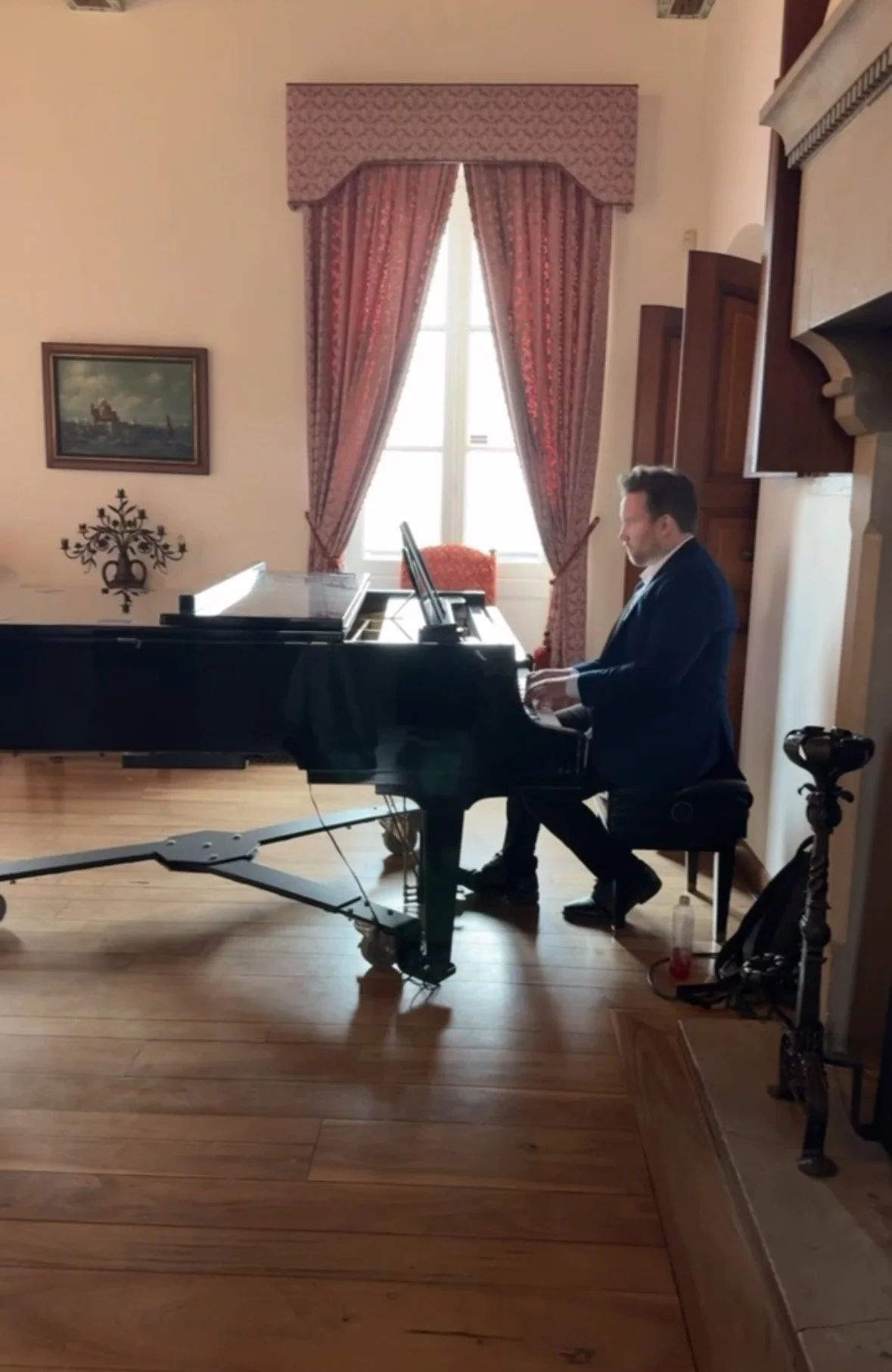 A man in a suit playing a grand piano in a room with wooden floors, pink curtains, and a framed painting on the wall.