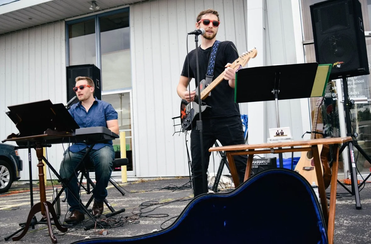 Two musicians performing outdoors in front of a white building, with one playing keyboard and singing into a microphone, and the other playing electric guitar and singing, both wearing sunglasses.