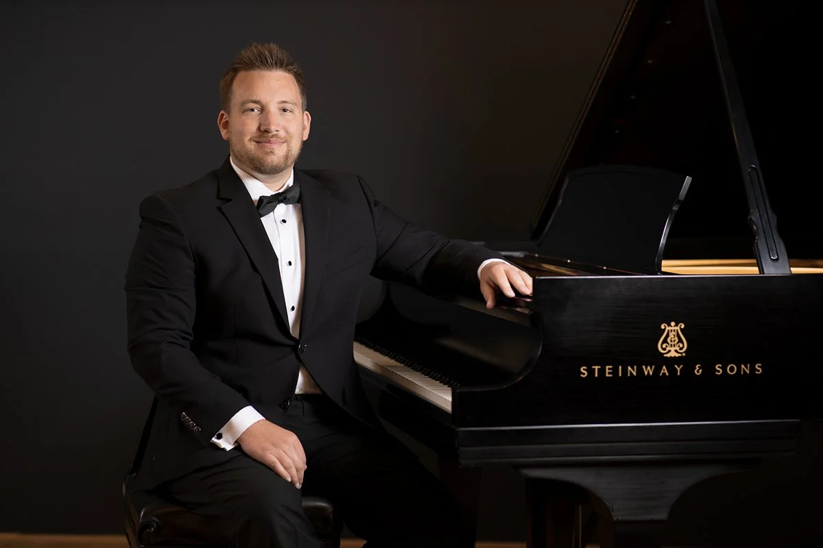 Man in formal tuxedo sitting at a black Steinway & Sons grand piano against a dark background.