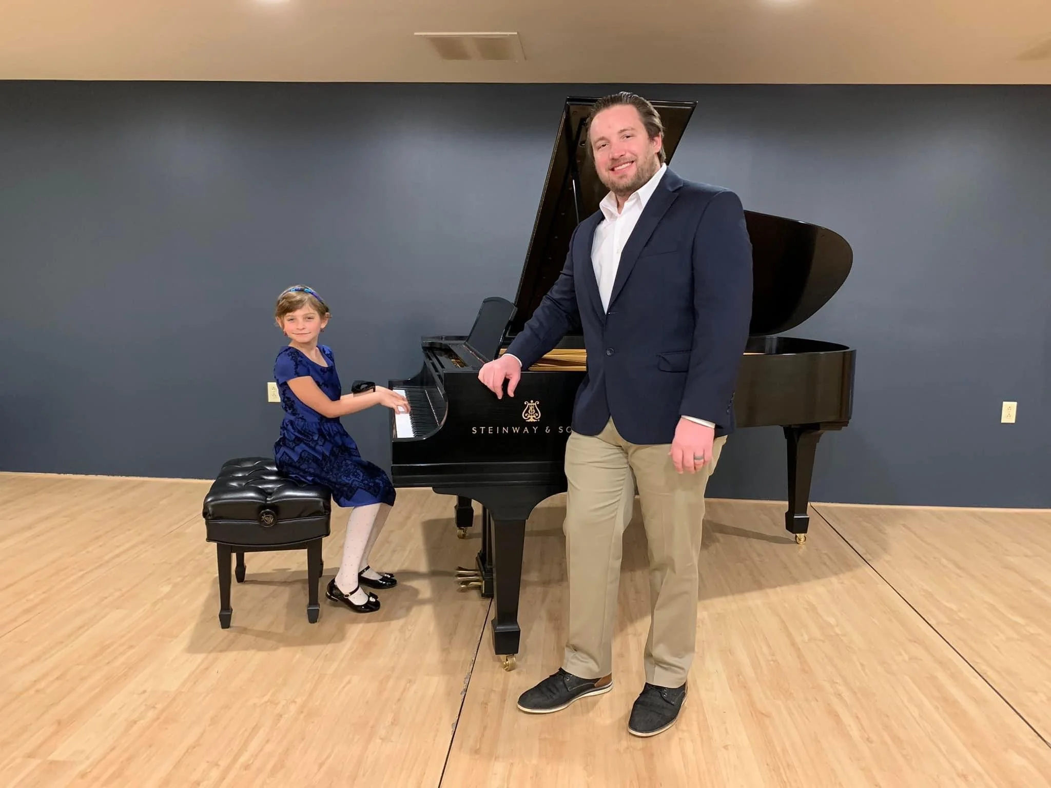A young girl in a blue dress and white tights playing a grand piano, with a man in a navy blazer and khaki pants standing beside them in a room with a blue wall and wooden floor.