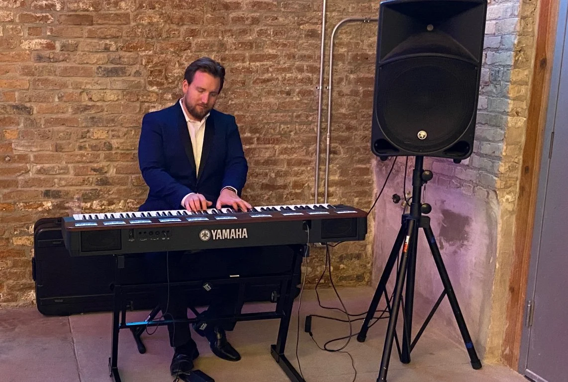 A man in a dark suit playing a Yamaha keyboard on a stand, with a large speaker on a tripod beside him, against a brick wall background.