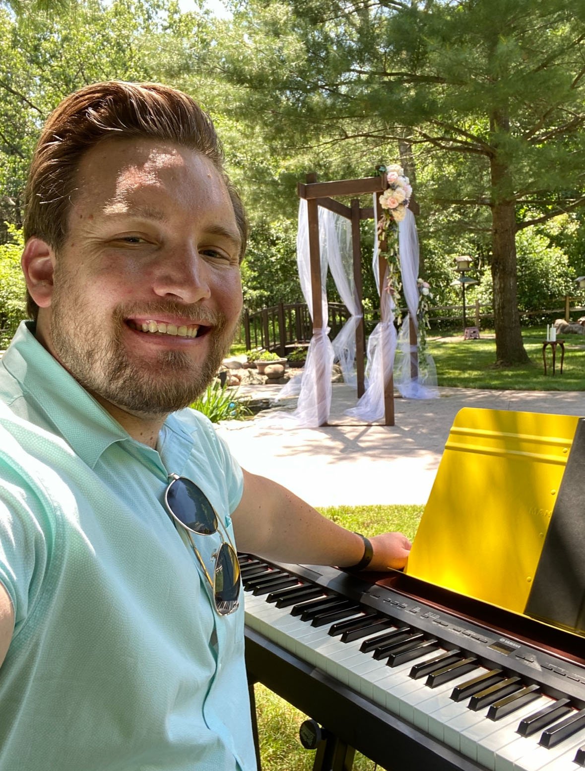 Man smiling at a keyboard piano outdoors near a wedding arch decorated with white fabric and flowers, in a lush green park setting.