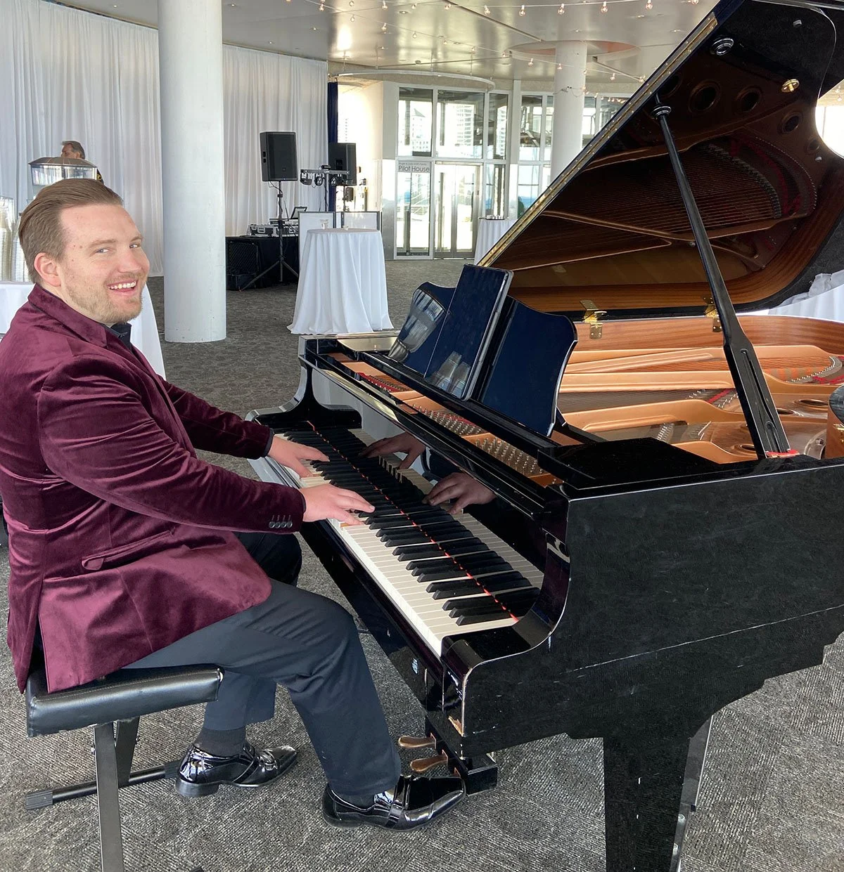 A man in a maroon velvet blazer playing a grand piano at an event space with white drapes and high windows.