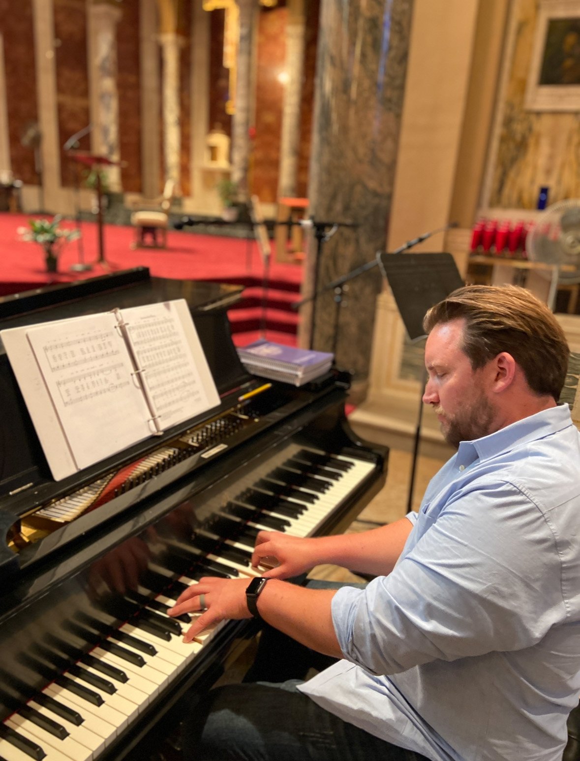 A man with light brown hair and a beard playing a black grand piano in a setting that appears to be a church or concert hall, with music sheets on the piano and a stage with a red carpet in the background.