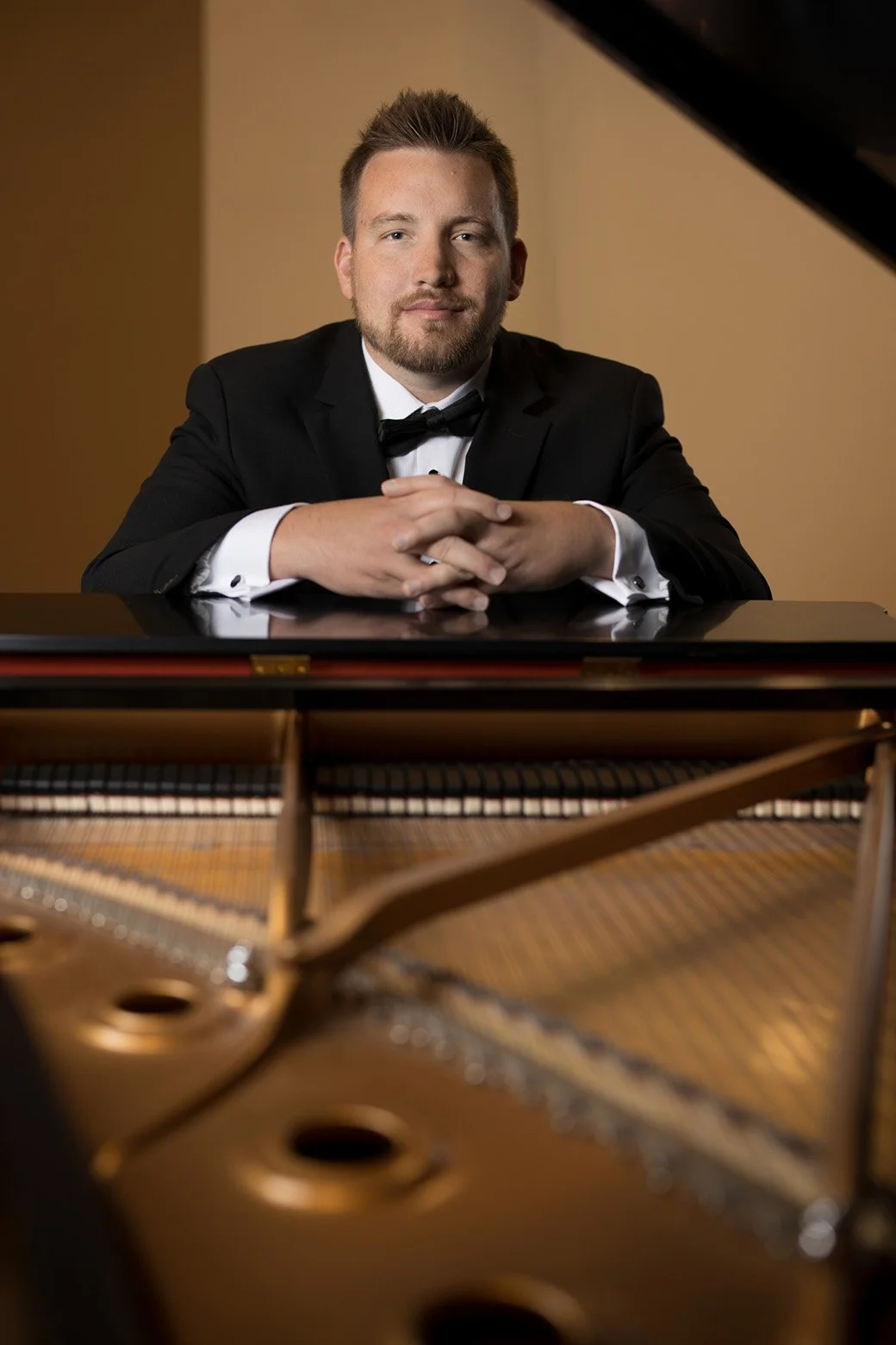 A man in a tuxedo sitting at a grand piano with his arms crossed, looking at the camera, with a beige wall in the background.