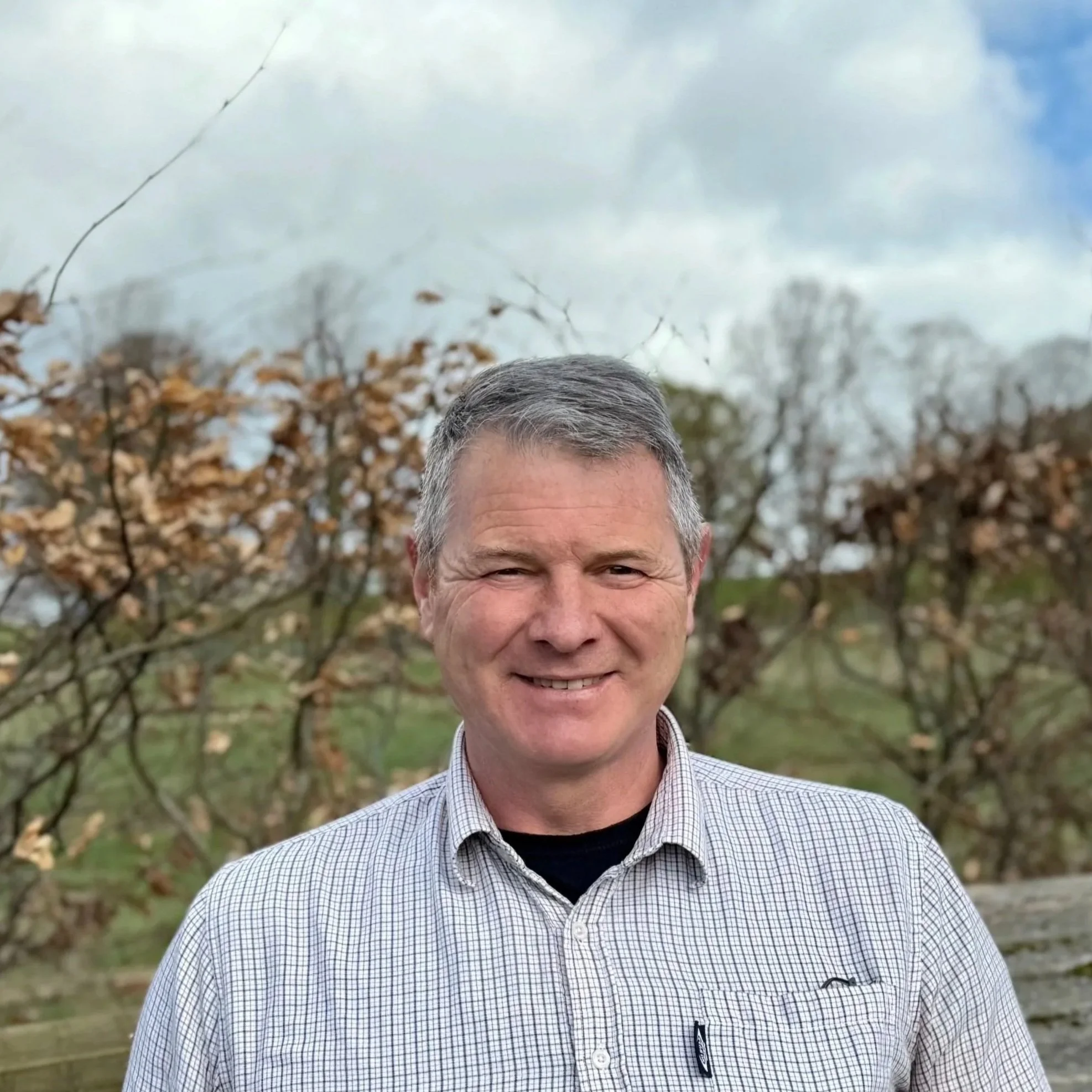 A smiling man with short gray hair wearing a checkered shirt standing outdoors in front of trees with dry leaves and a partly cloudy sky.