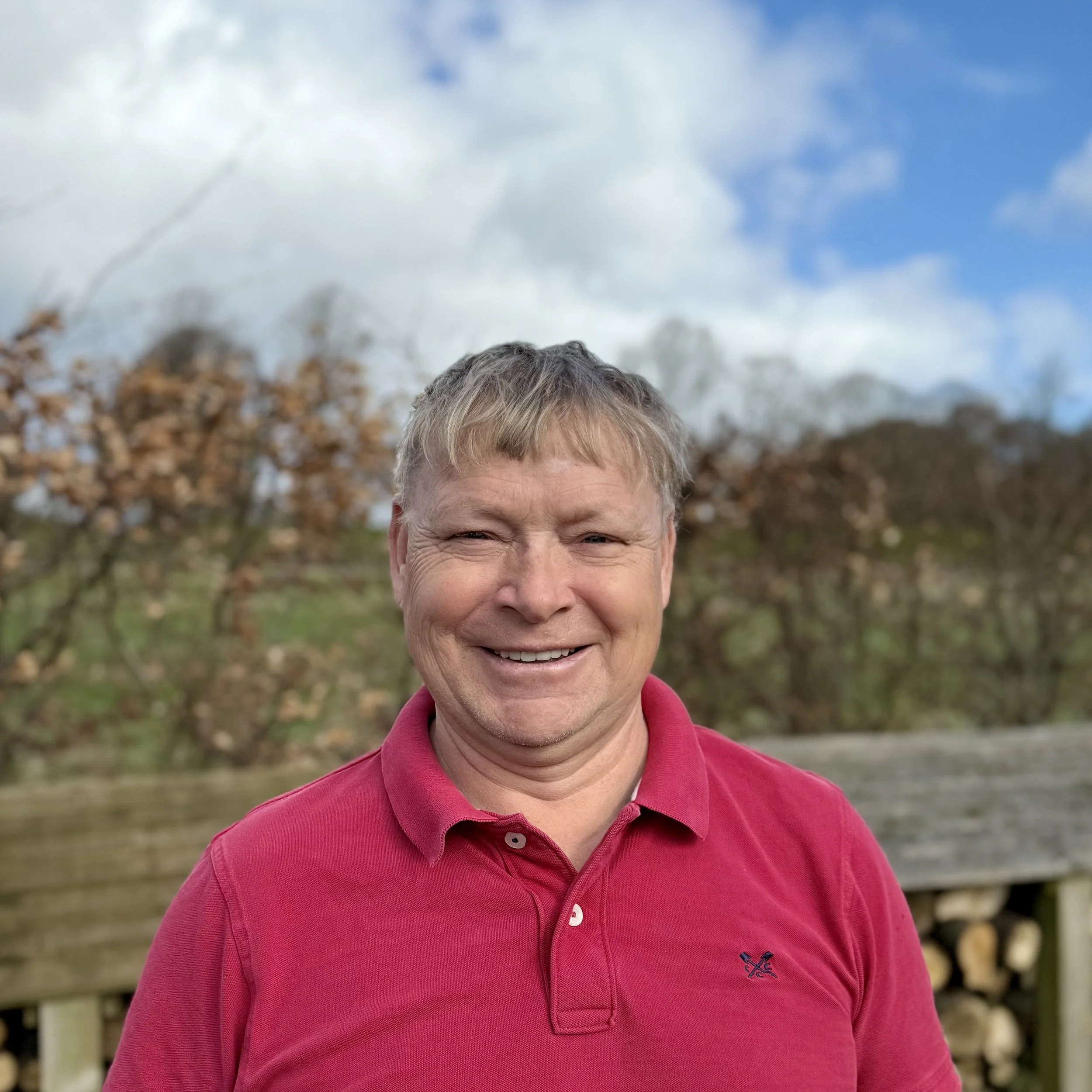 A smiling man in a red polo shirt outdoors with trees and a wooden fence in the background under a partly cloudy sky.