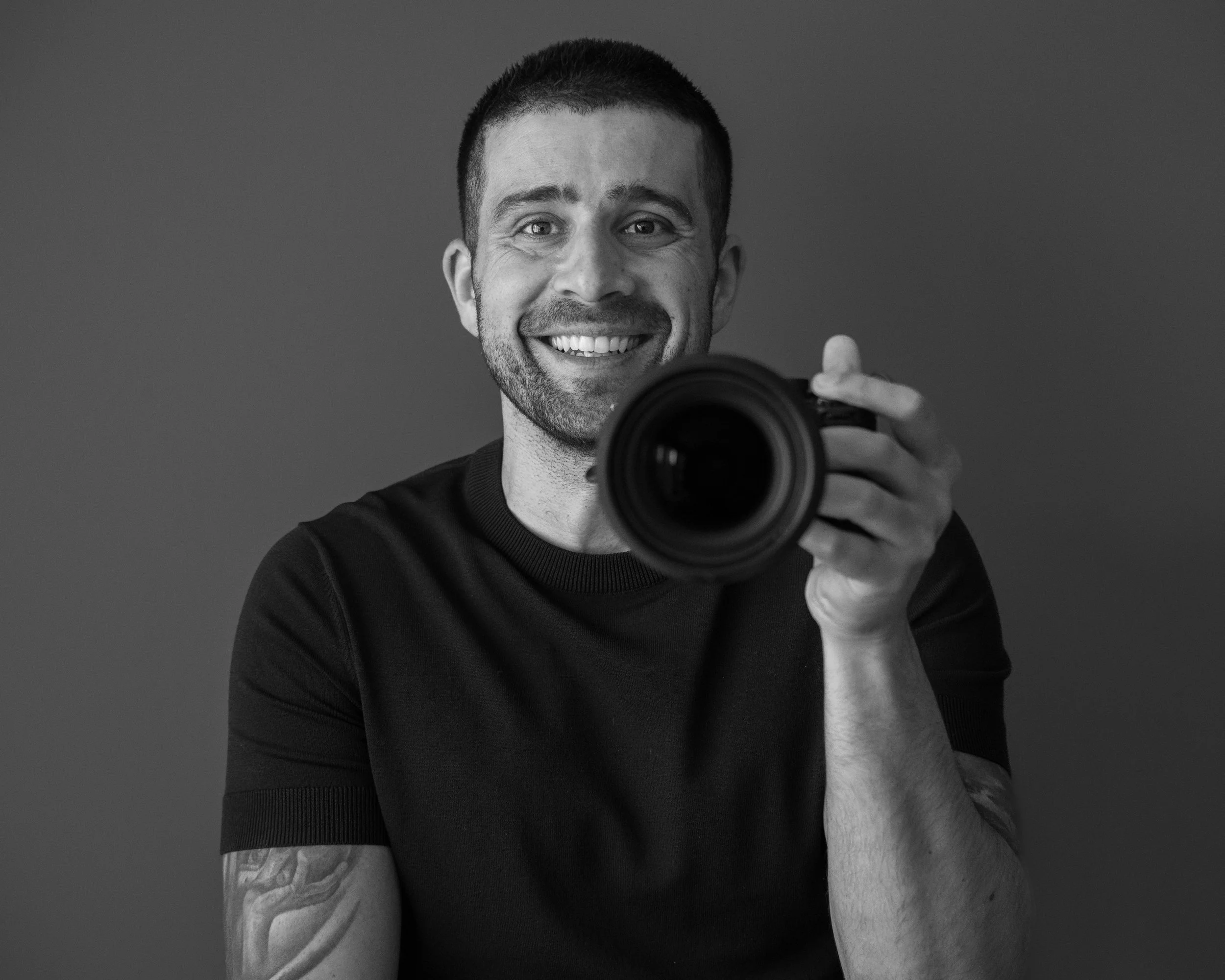 A smiling man with short dark hair and a beard holding a camera, taking a selfie in front of a plain background.