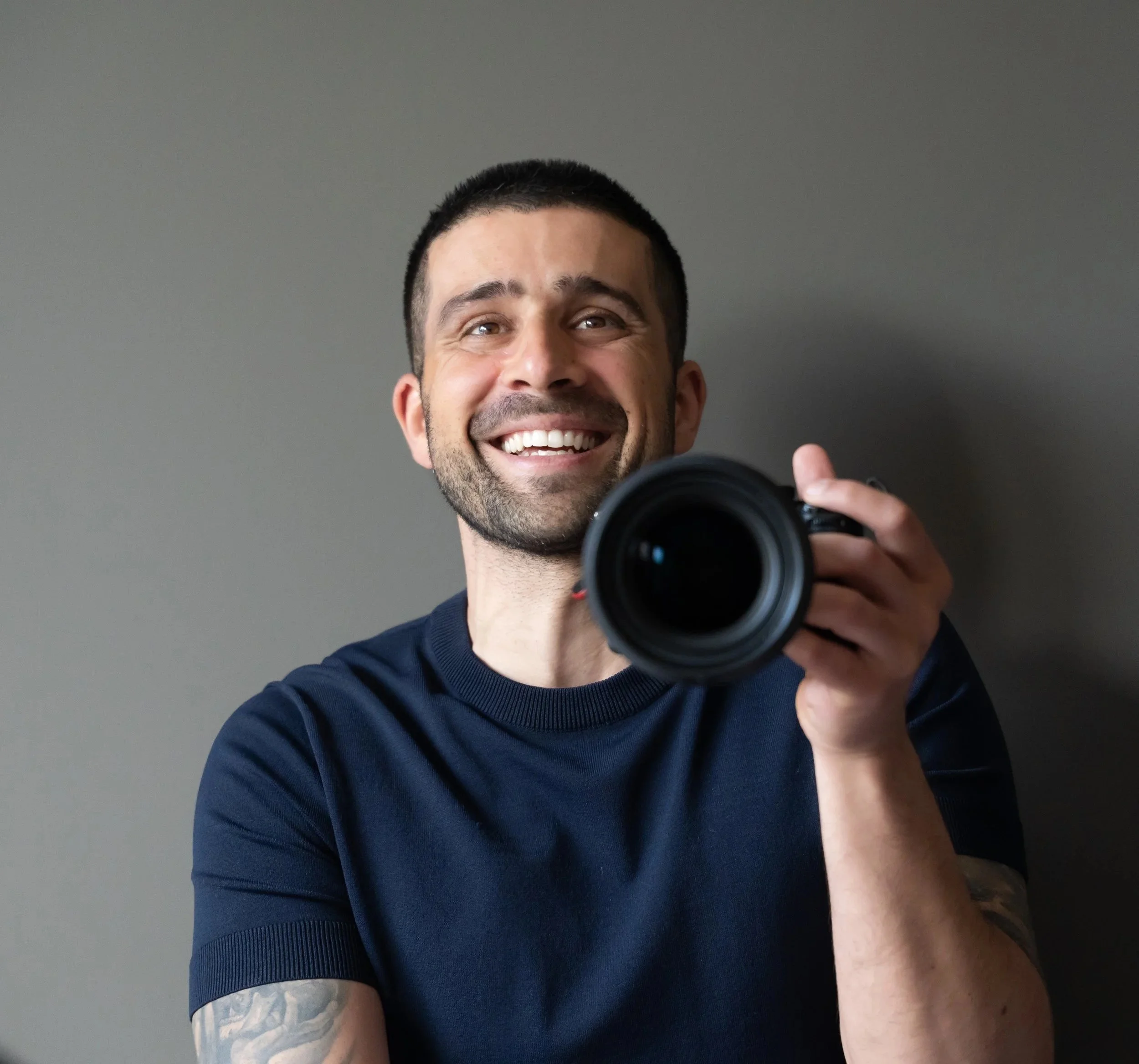Man with dark hair and beard smiling while taking a selfie with a camera, wearing a navy blue shirt against a gray wall.