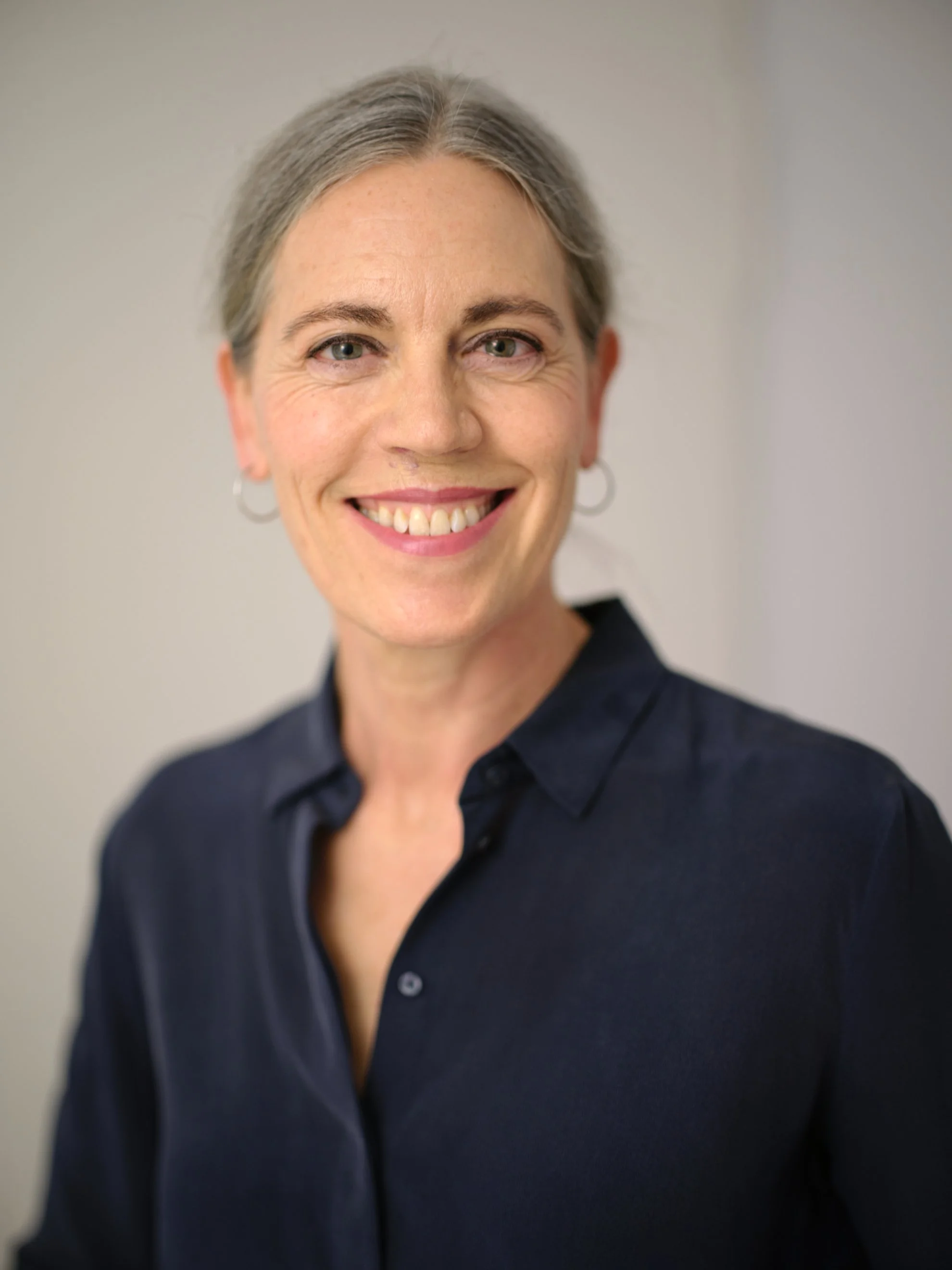 Nahaufnahme einer lachenden Frau mit kurzen grauen Haaren, blauen Augen, blauser Bluse und Ohrhaken, vor neutralem Hintergrund.