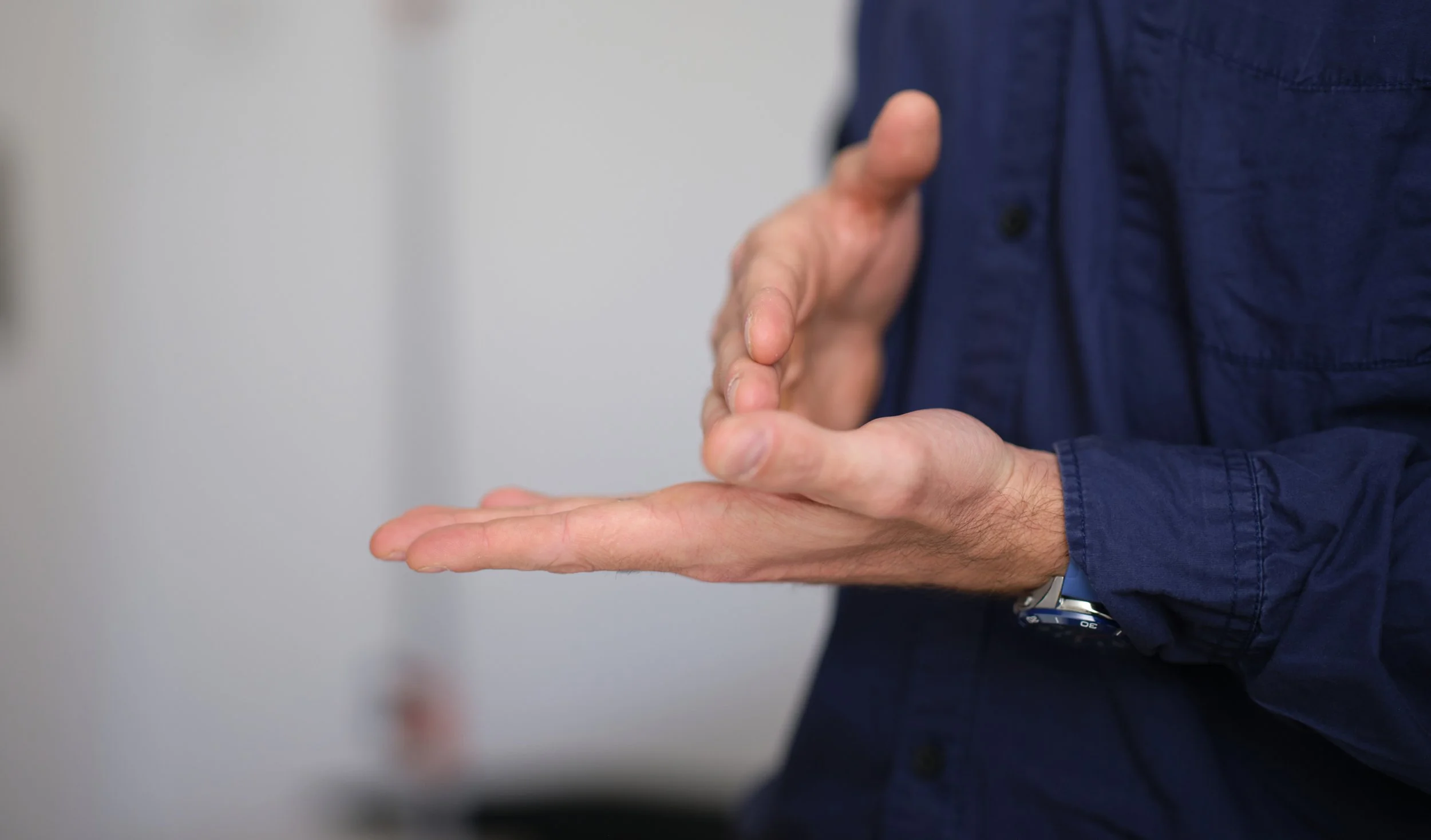 Hände einer Person in dunklem Hemd, die eine Geste macht, bei der eine Hand flach auf den Handrücken der anderen Hand ruht.