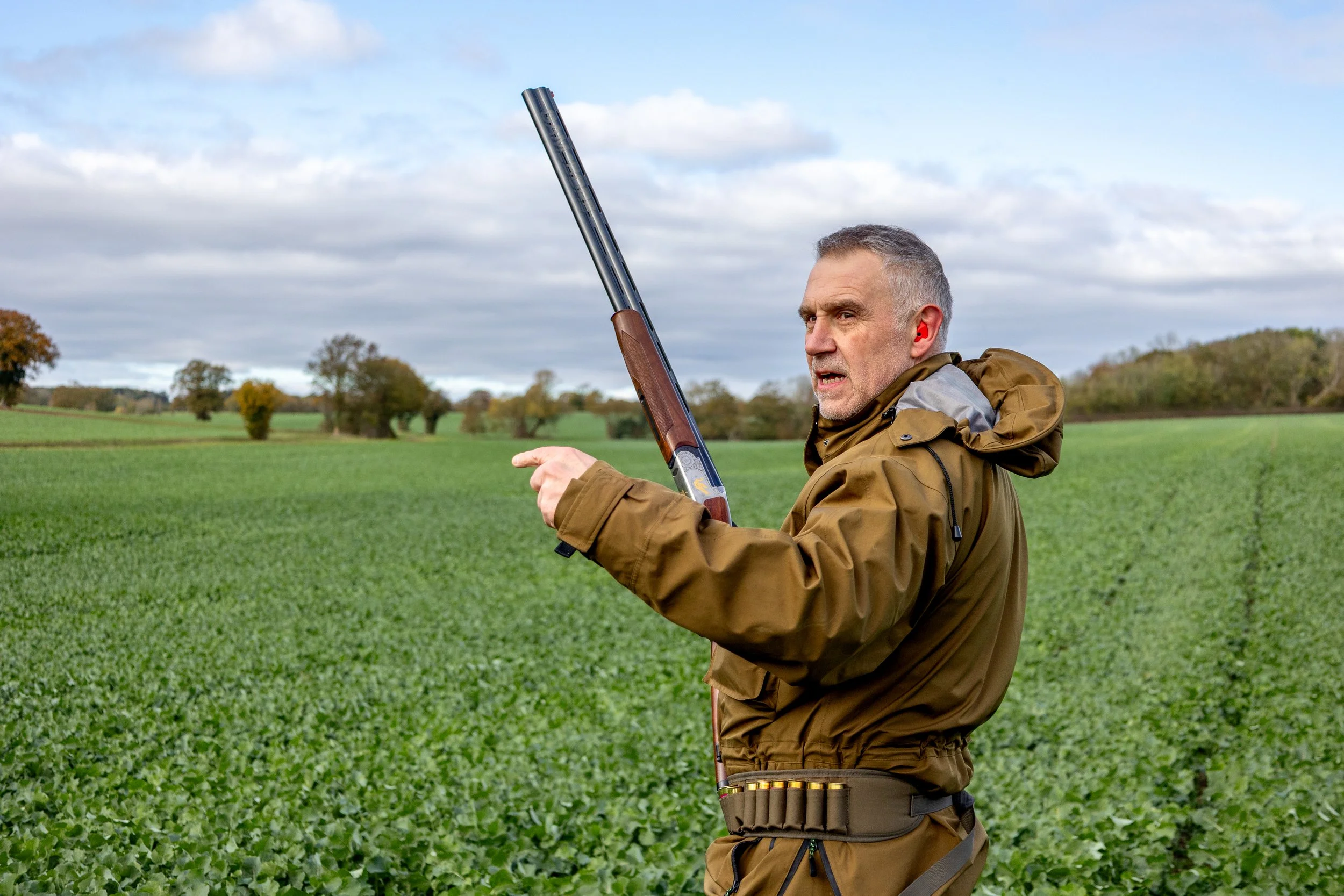 A middle-aged man with gray hair, wearing a brown jacket and ear protection, holding a shotgun while pheasant shooting, pointing and speaking outdoors in a green field with trees under a cloudy sky.