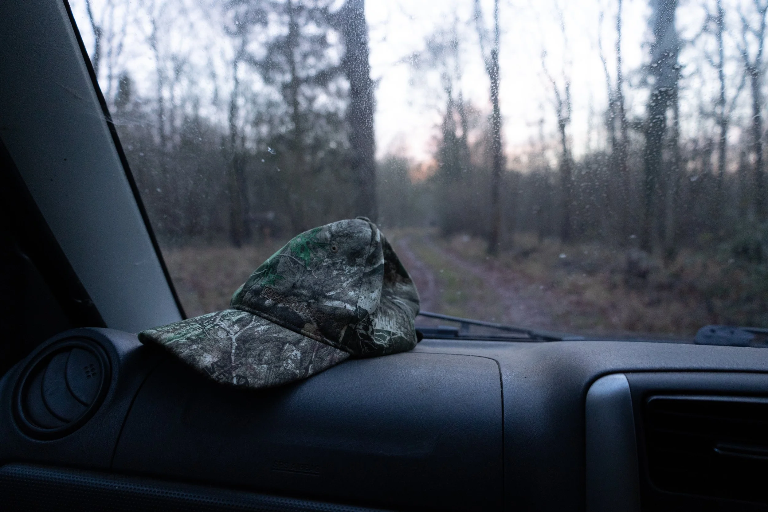 Camouflage baseball cap on the dashboard of a vehicle, with a wooded dirt road visible through a wet windshield.