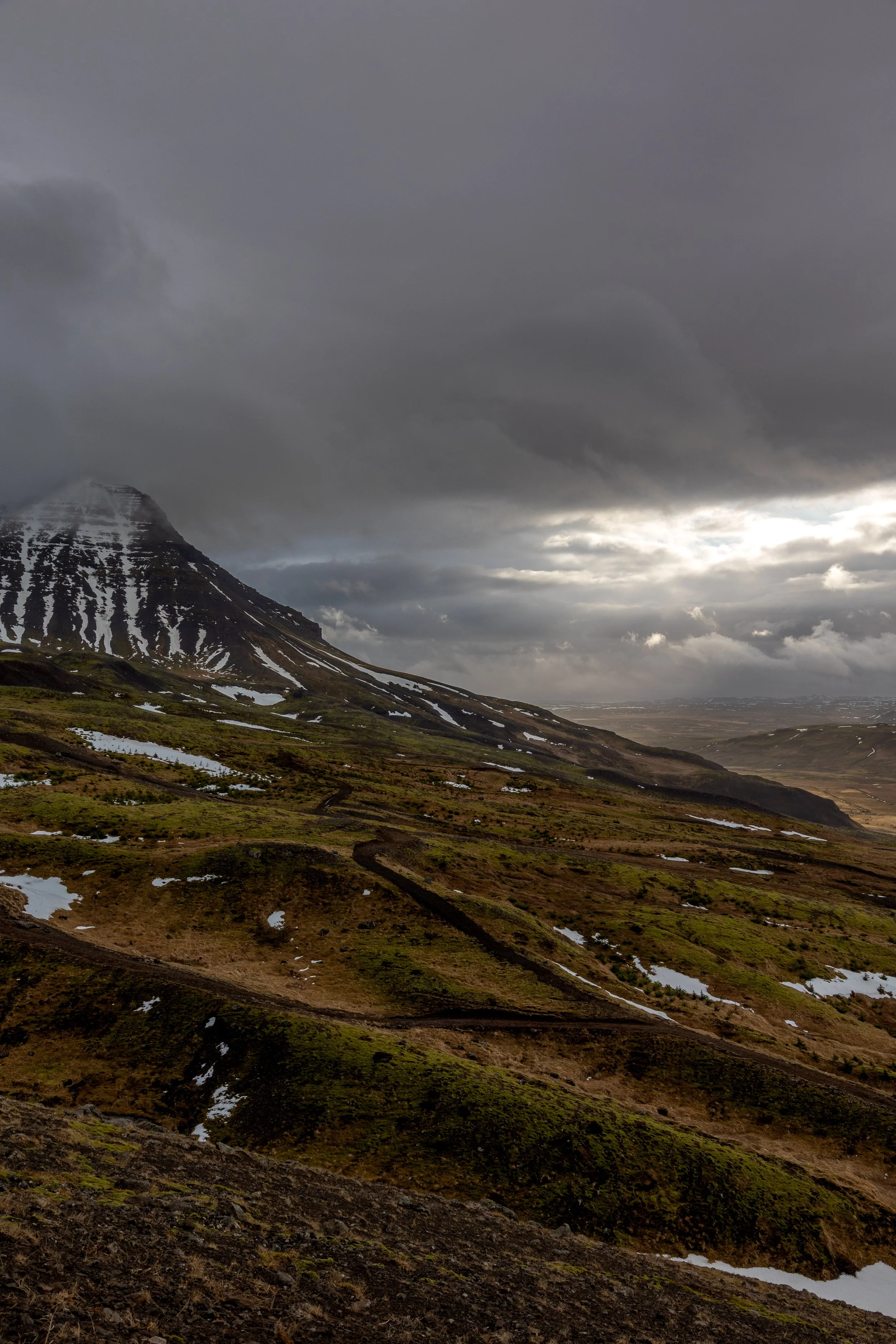 A mountainous landscape under a dark, cloudy sky with patches of sunlight breaking through, snow on the mountain peak, and winding dirt roads across green and brown terrain.
