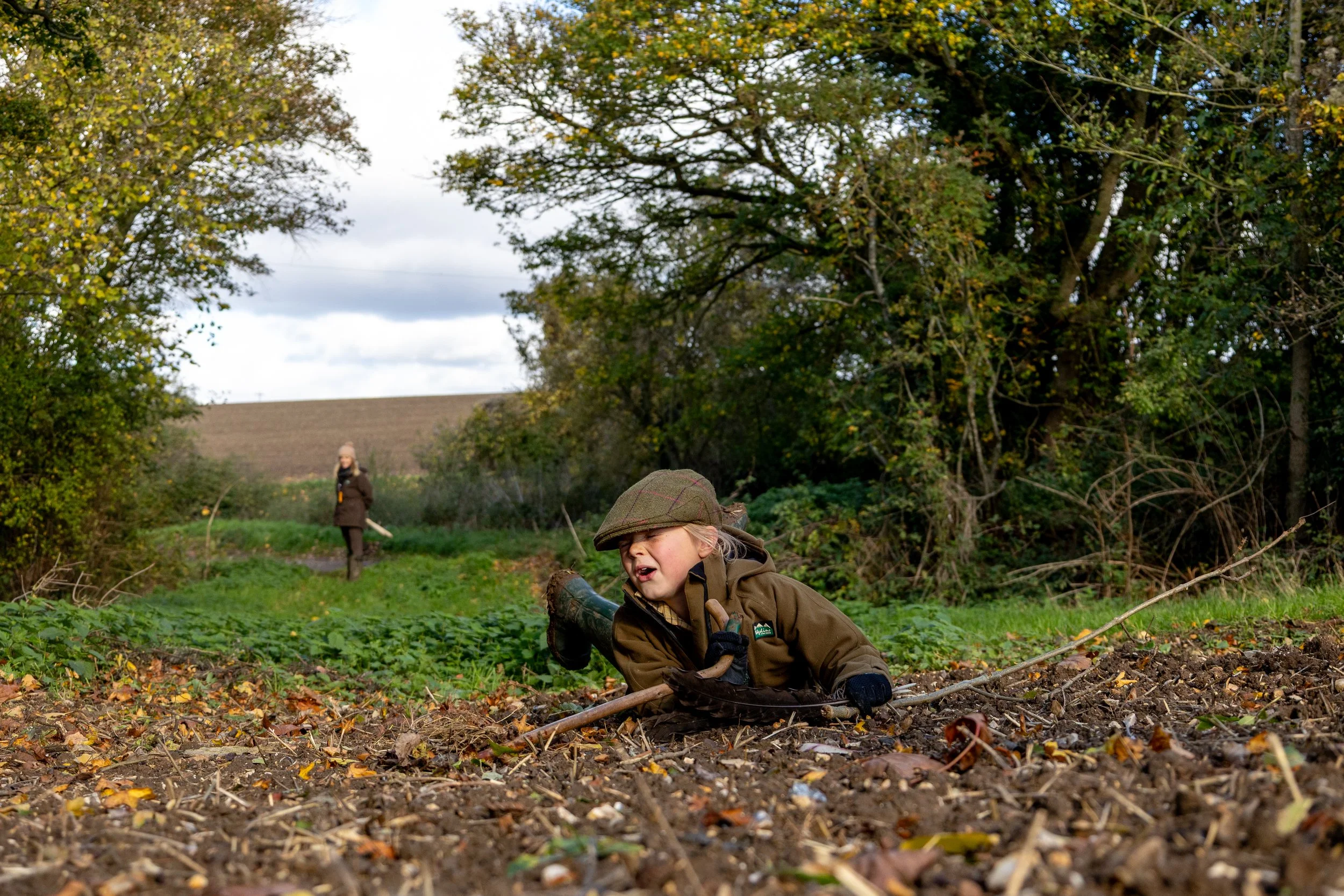 A young child wearing outdoor clothing and a cap is crawling on the ground with a stick in hand, in a forested area during autumn, with another person walking in the background.