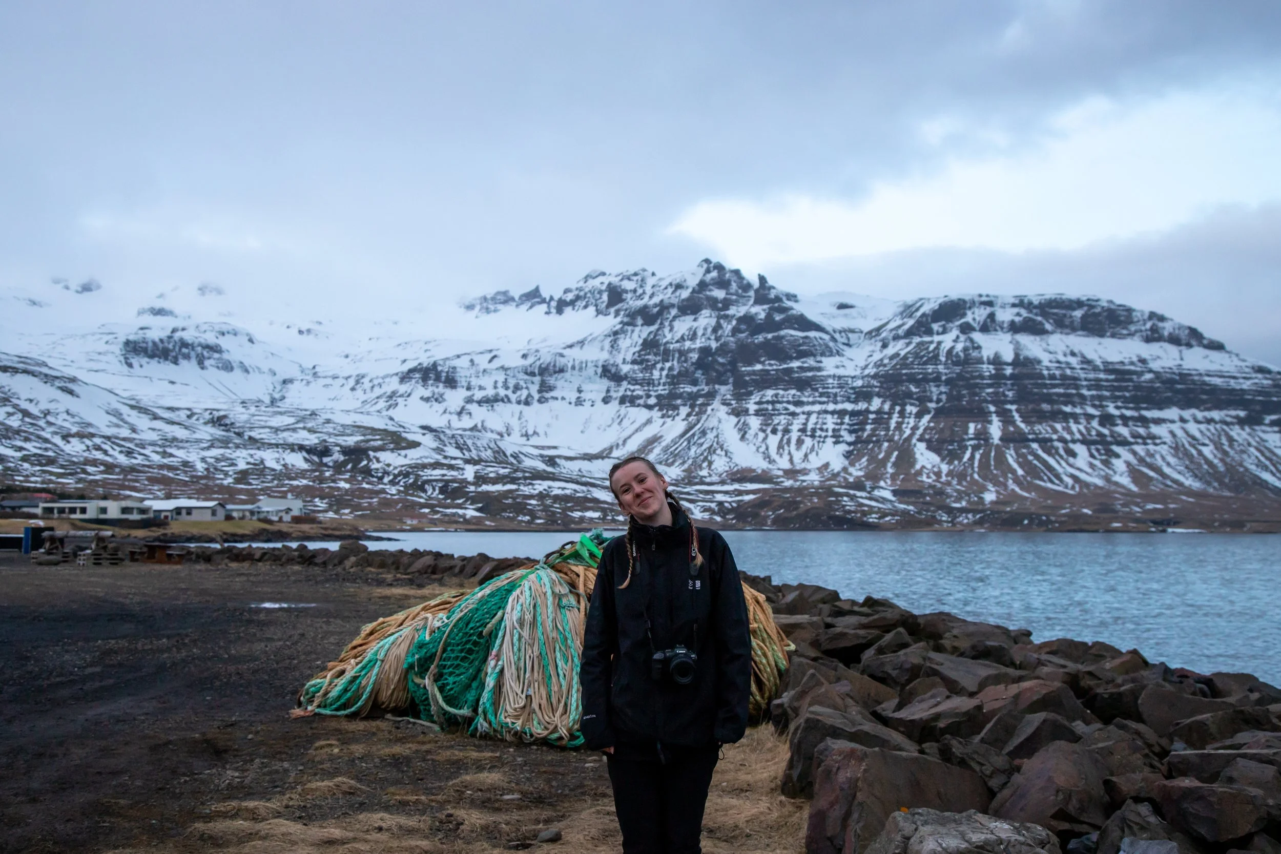A woman photographer dressed in black outdoor waterproof warm clothing standing on a rocky shoreline in Iceland with large fishing nets behind her, a lake, snowy mountains, and an overcast sky in the background.