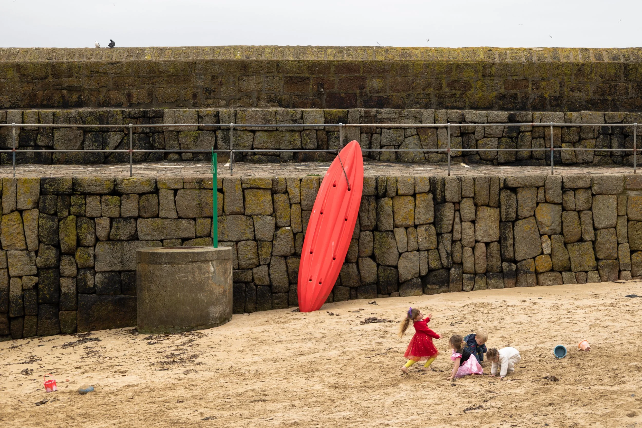Mousehole, Cornwall. Girl in red dress with red kayak on beach with beach wall