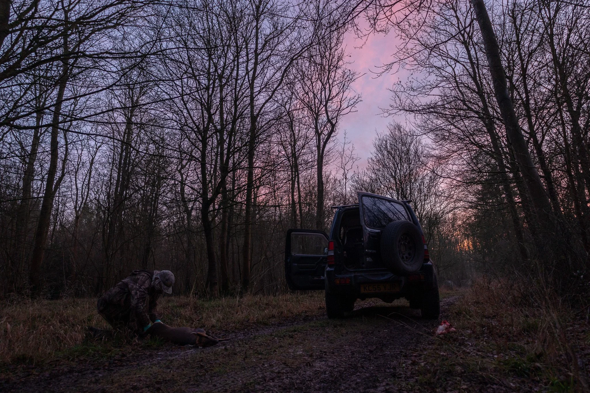 A person wearing camouflage clothing and a cap is kneeling next to a deer on the ground in a wooded area at dusk. An open vehicle with a spare tire mounted on the back is parked nearby on a dirt path.