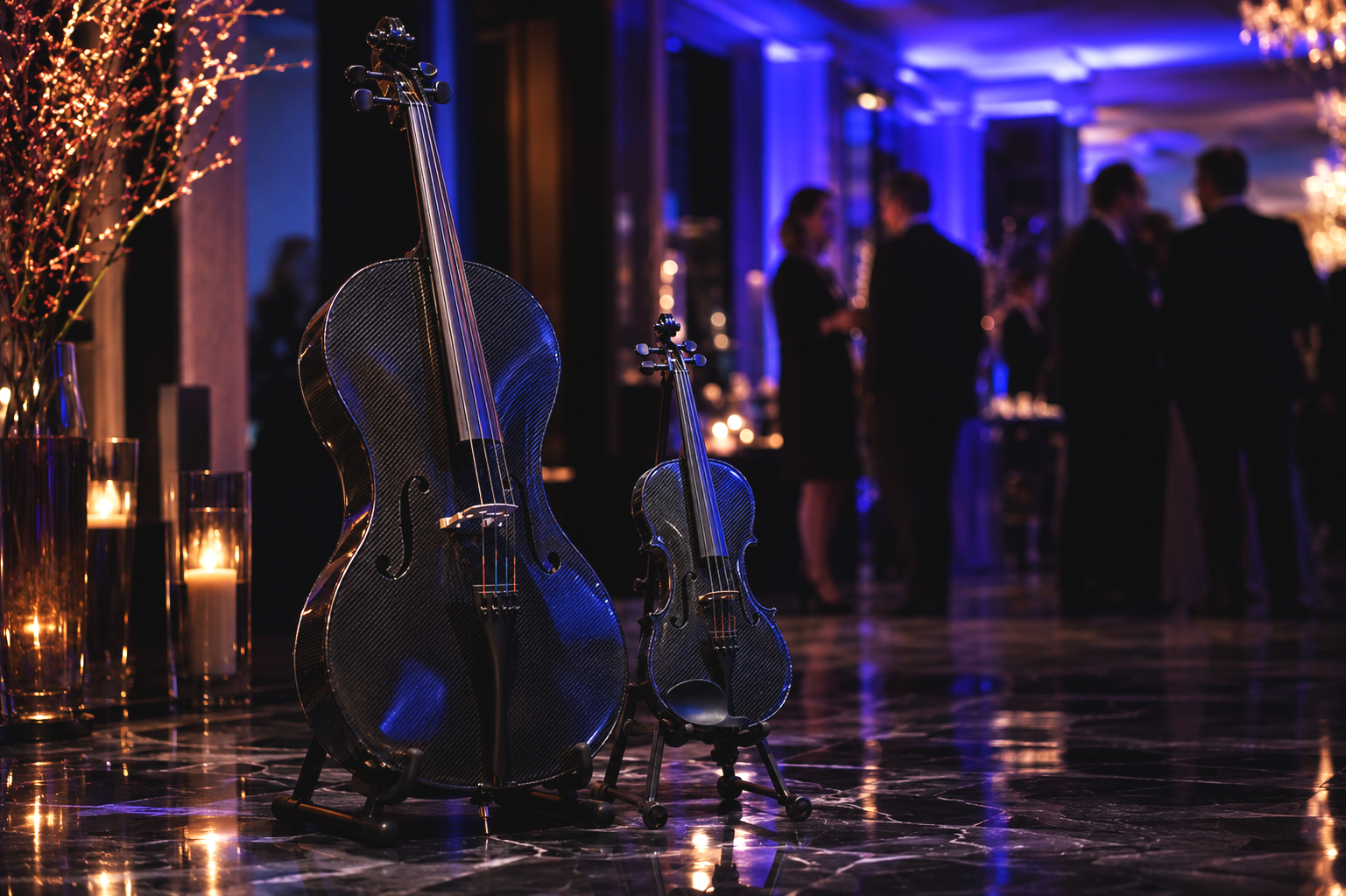 Two carbon fiber violins, one large and one small, are displayed on stands in a dimly lit, elegant indoor setting with glowing candles and blurred people in the background, suggesting a formal event or concert.