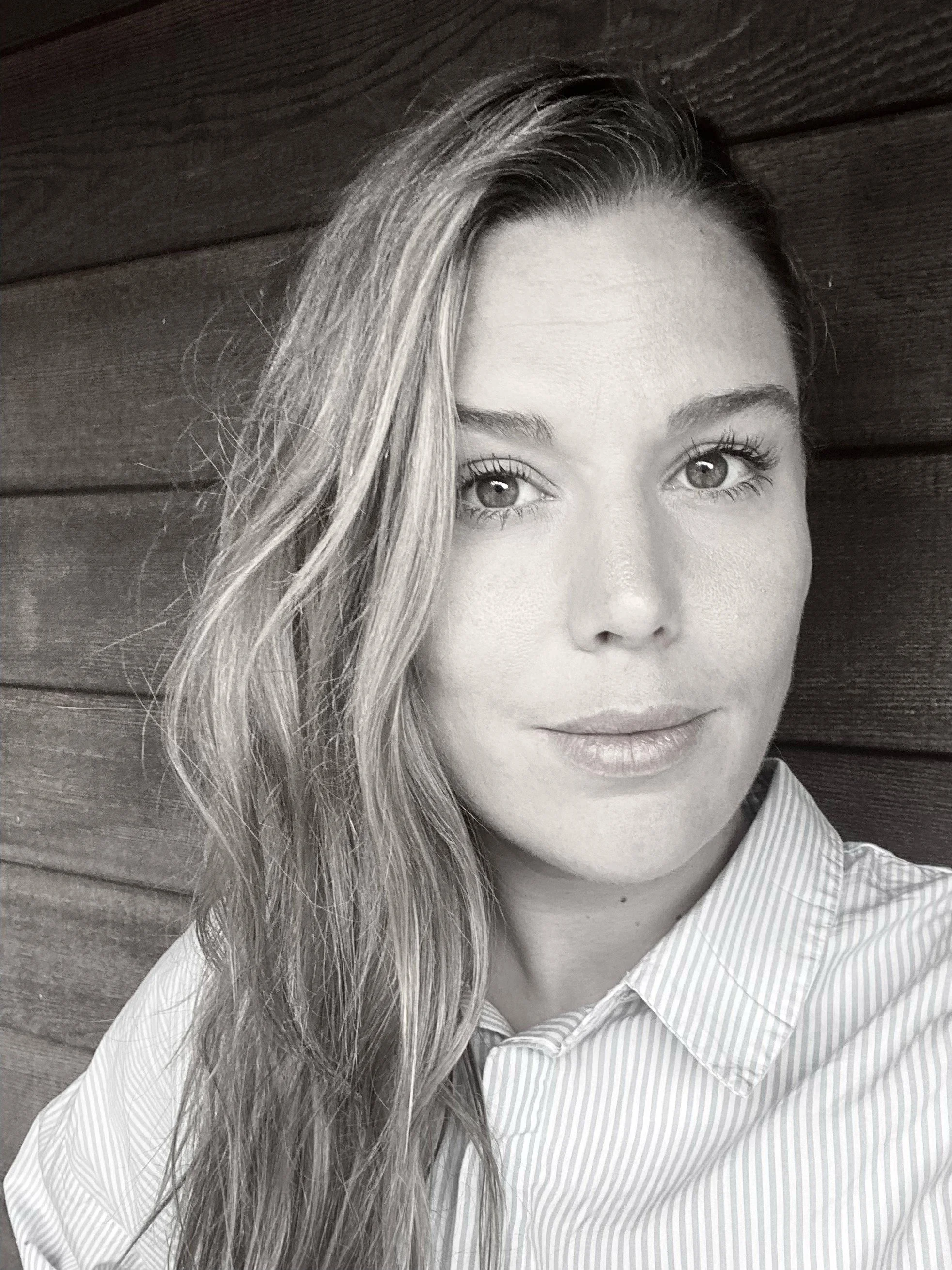 A black-and-white portrait of a woman with long, wavy hair, light eyes, and a slight smile, wearing a striped collared shirt, posed against a wooden wall background.