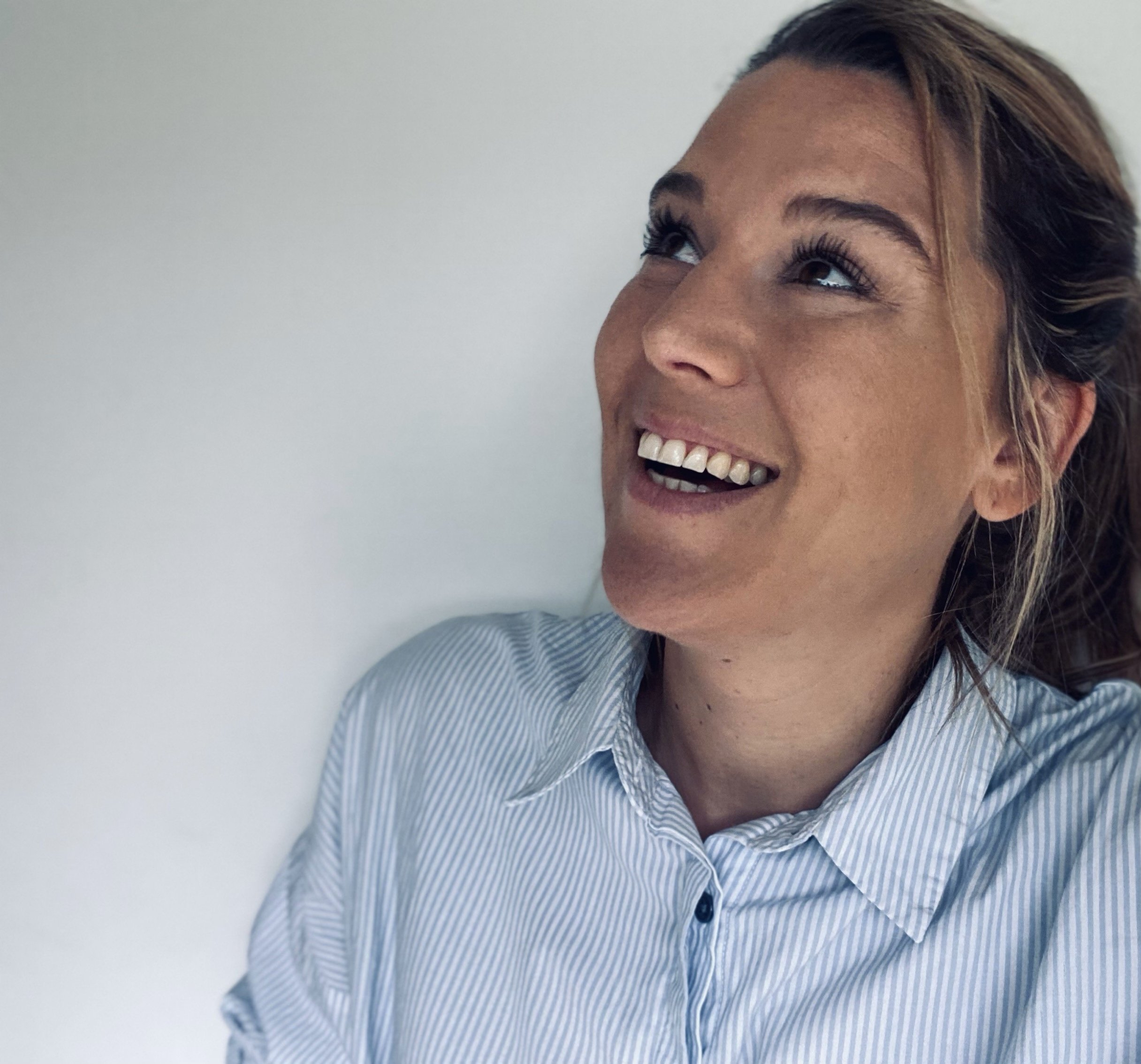A woman with brown hair wearing a light blue and white striped shirt, smiling and looking upwards against a plain white background.