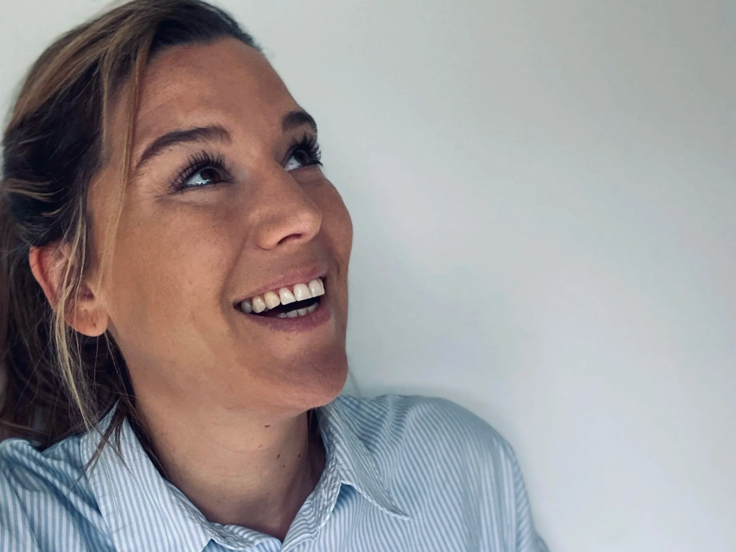 A woman with light brown hair and blue eyes is smiling and looking upward. She is wearing a light blue striped collared shirt and is standing against a plain light-colored wall.
