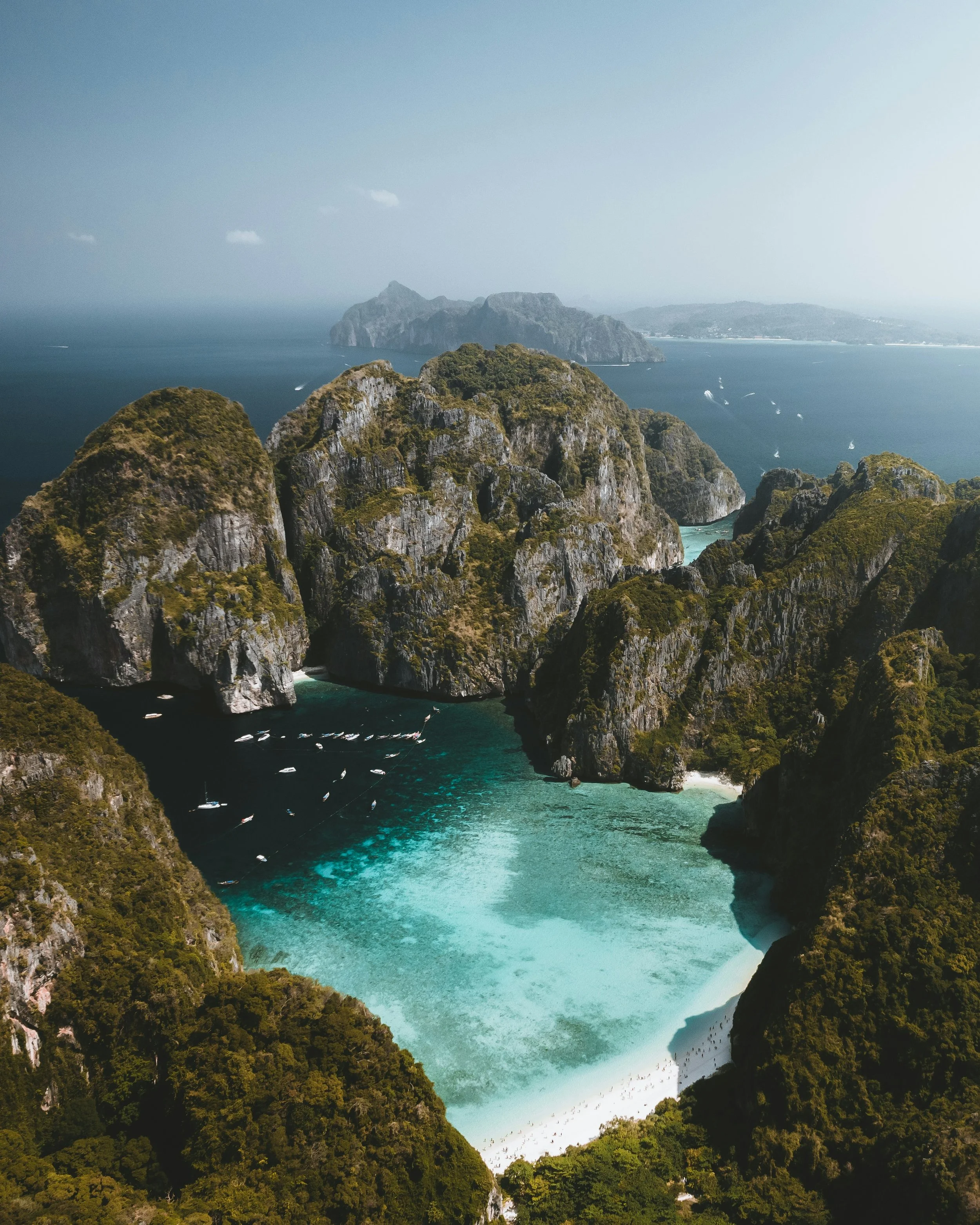 Paysage côtier avec des formations rocheuses, une plage de sable blanc et un lagon turquoise entourés de végétation dense, avec la mer et d'autres îles au loin.