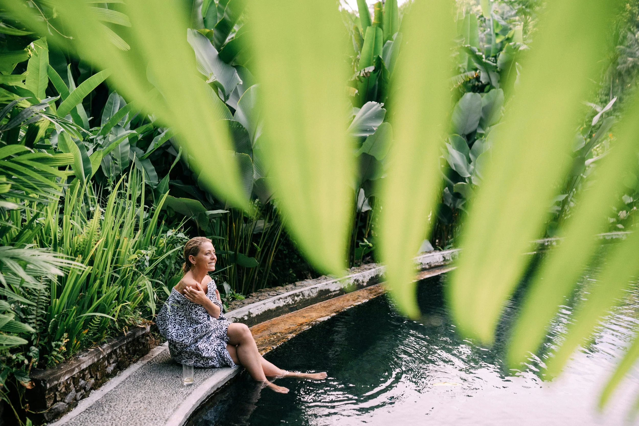 Femme assise au bord d'une eau noire entourée de végétation luxuriante, avec de grandes feuilles vertes en avant-plan.