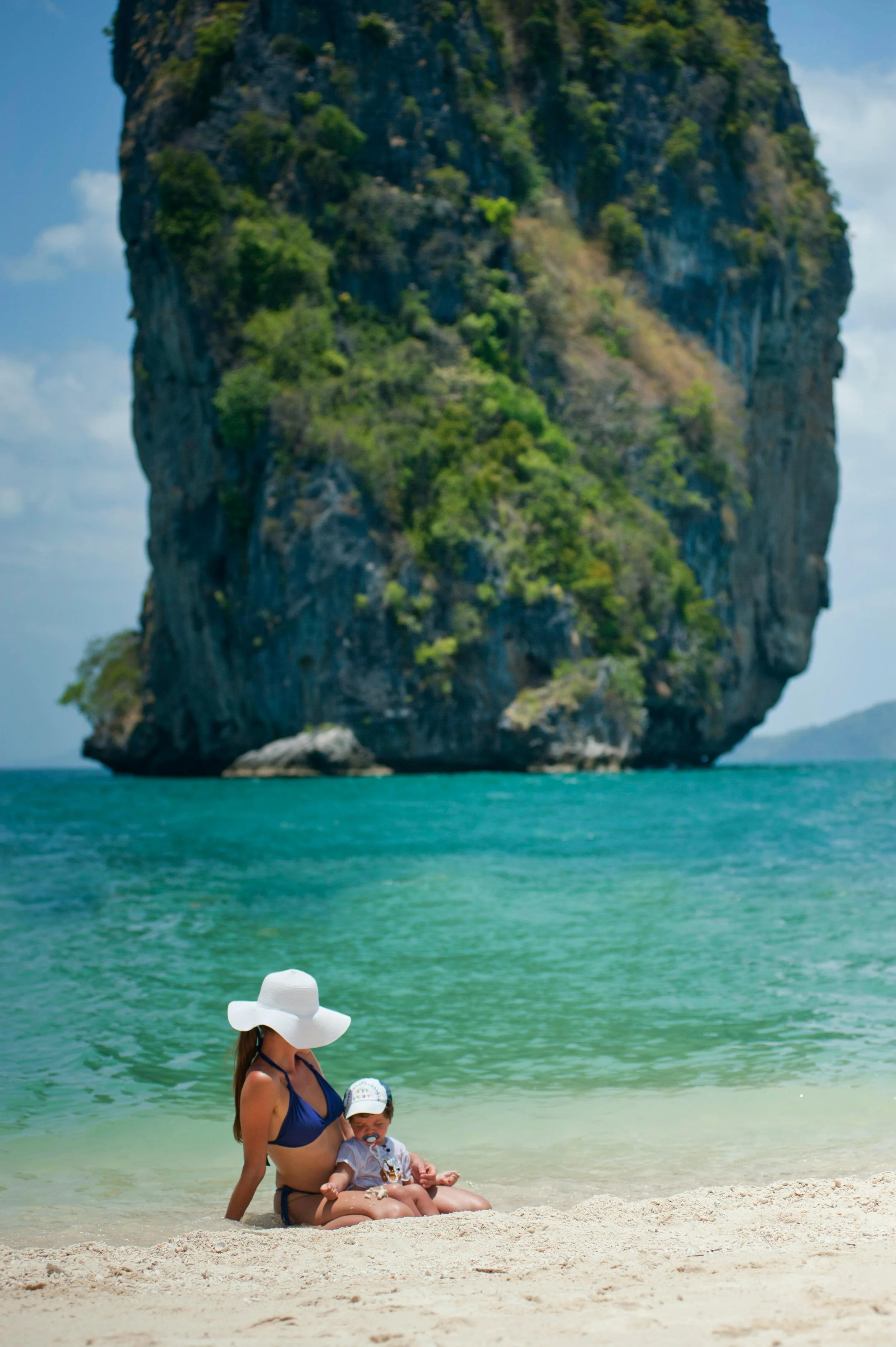 Une femme avec un chapeau blanc et un maillot de bain bleu assise sur la plage avec un bébé, devant une mer turquoise et un rocher recouvert de végétation.