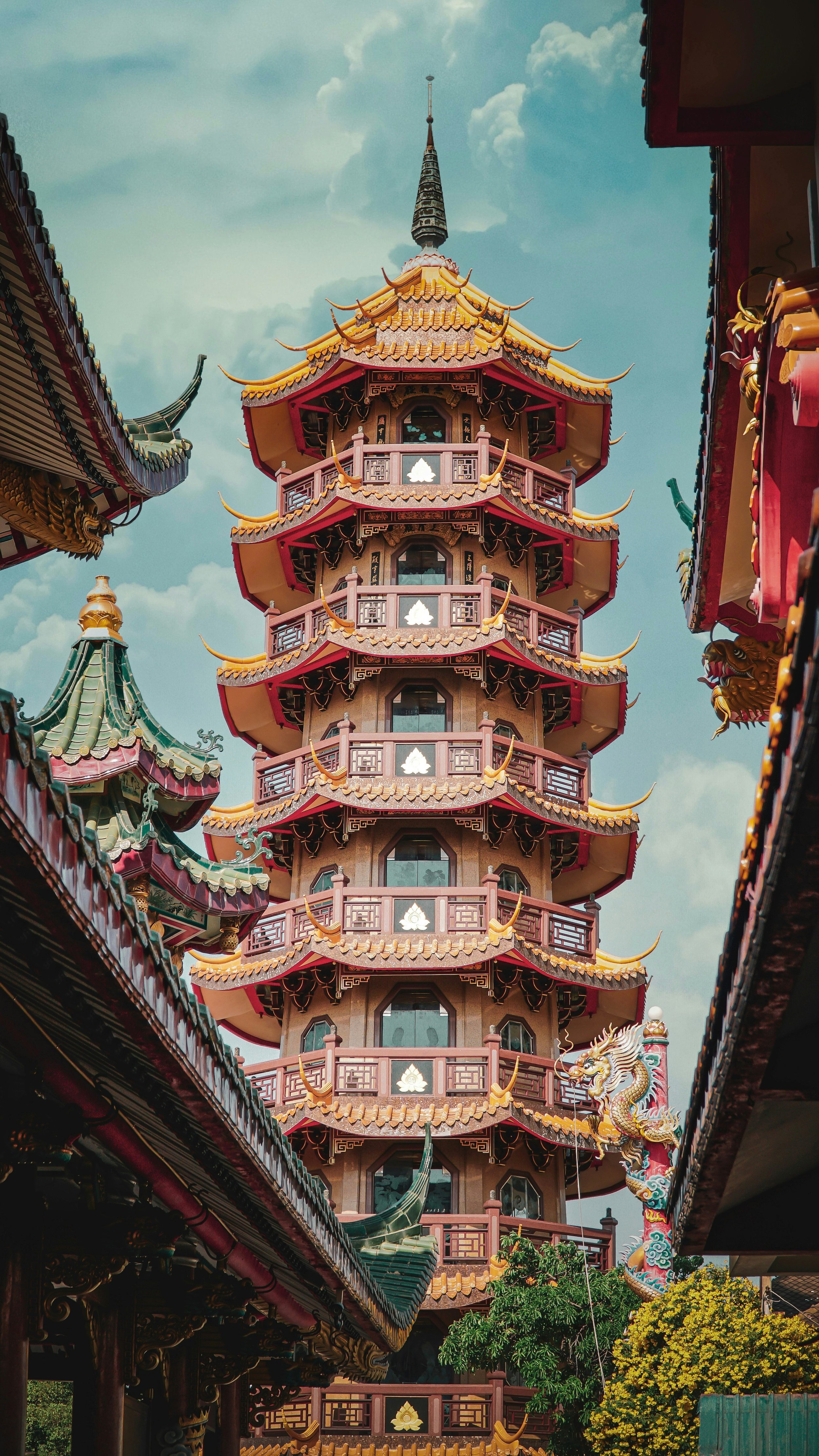 Une pagode traditionnelle à plusieurs étages avec des toits courbes en tuiles orange et des décorations dorées, entourée de bâtiments de style asiatique avec des toits verts, dans un environnement lumineux avec un ciel partiellement nuageux.