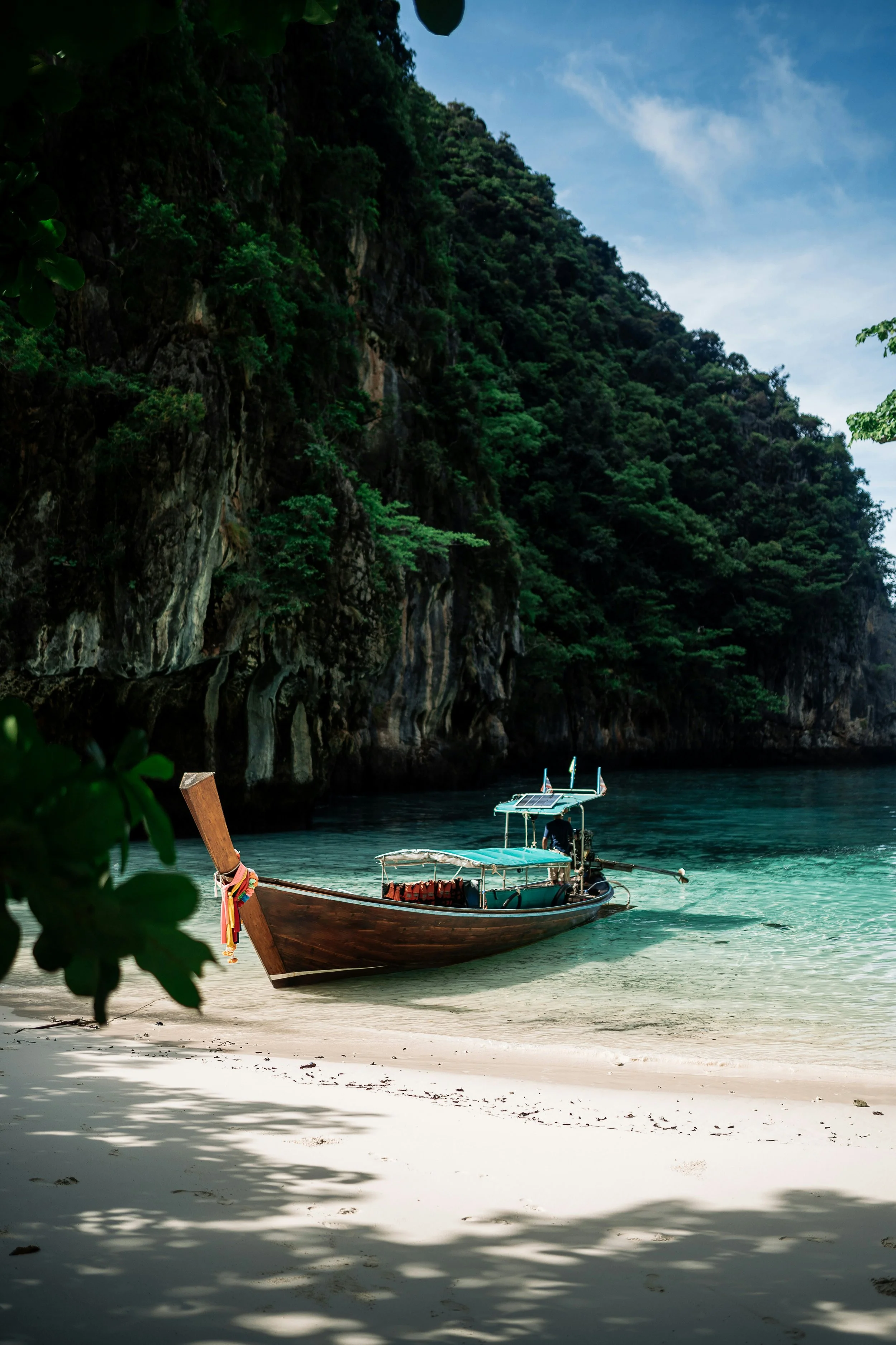 Une bateau traditionnel en bois repose sur une plage de sable blanc, avec des eaux turquoise et une falaise rocheuse verdoyante en arrière-plan.