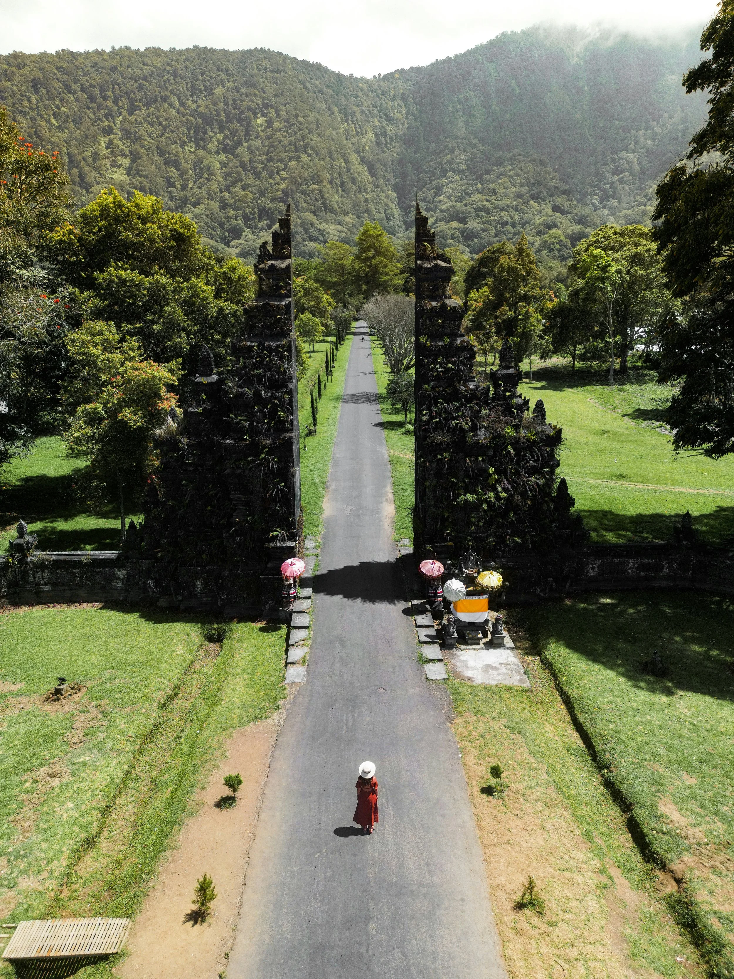 Une femme portant un chapeau blanc marche sur une route à l'entrée d'un temple avec des portes décoratives, entouré de nature verdoyante et de montagnes en arrière-plan.