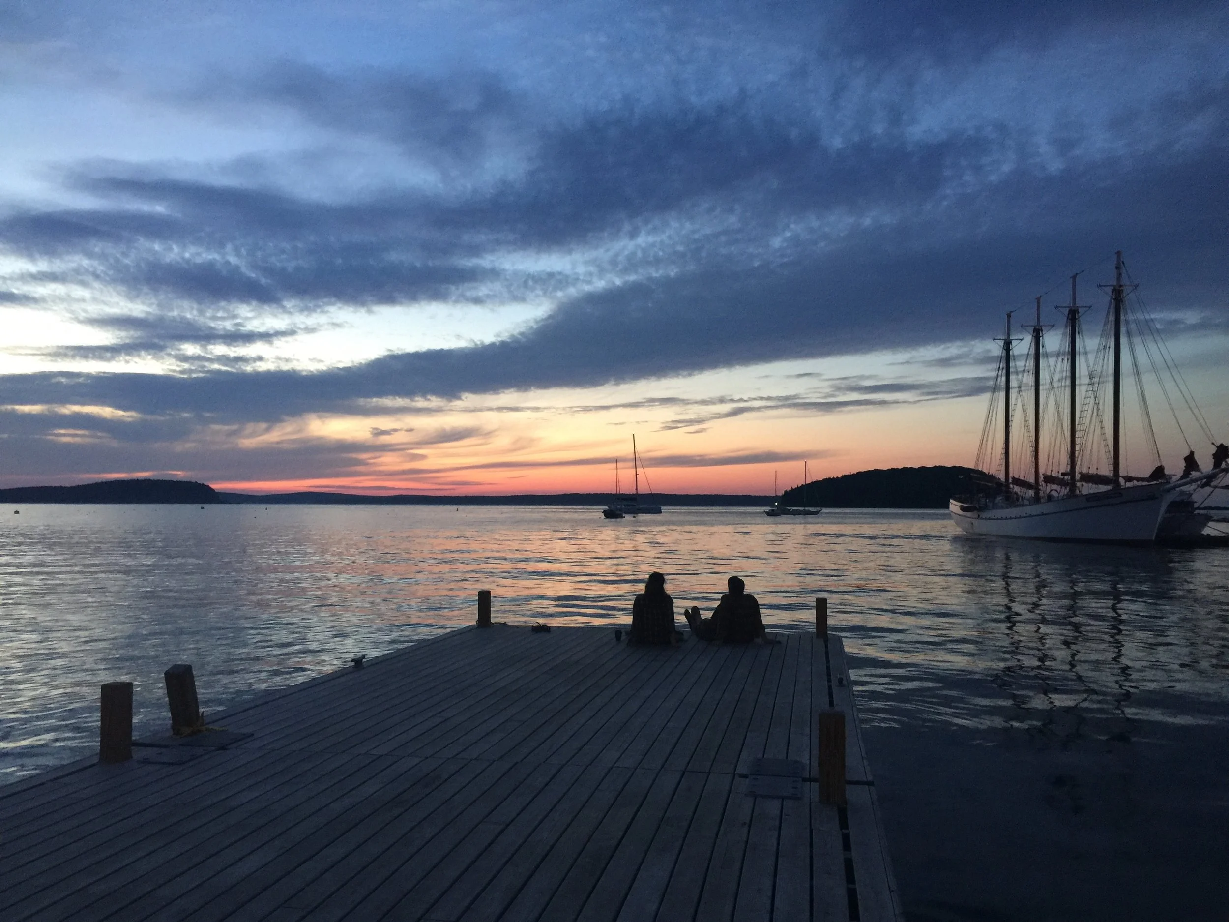 Couple sitting on a dock at sunset, overlooking calm water with sailboats docked and a distant hill in the background.