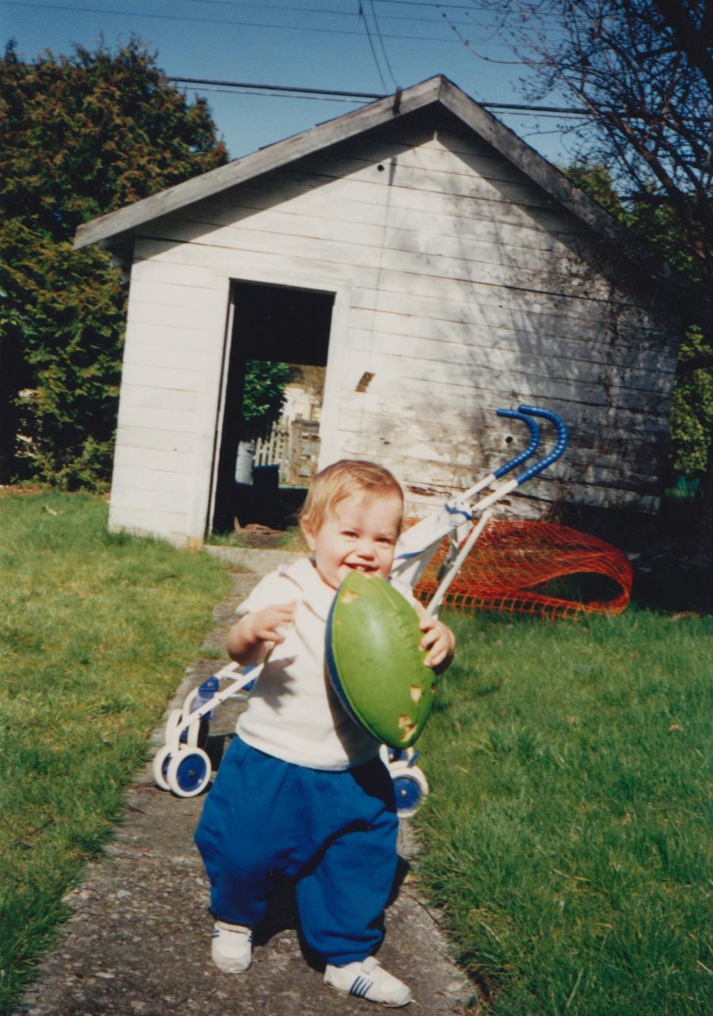 A young child smiling and playing outside with a green frisbee in hand, standing on a small paved path in front of a wooden shed with a grass yard and a toy stroller nearby.