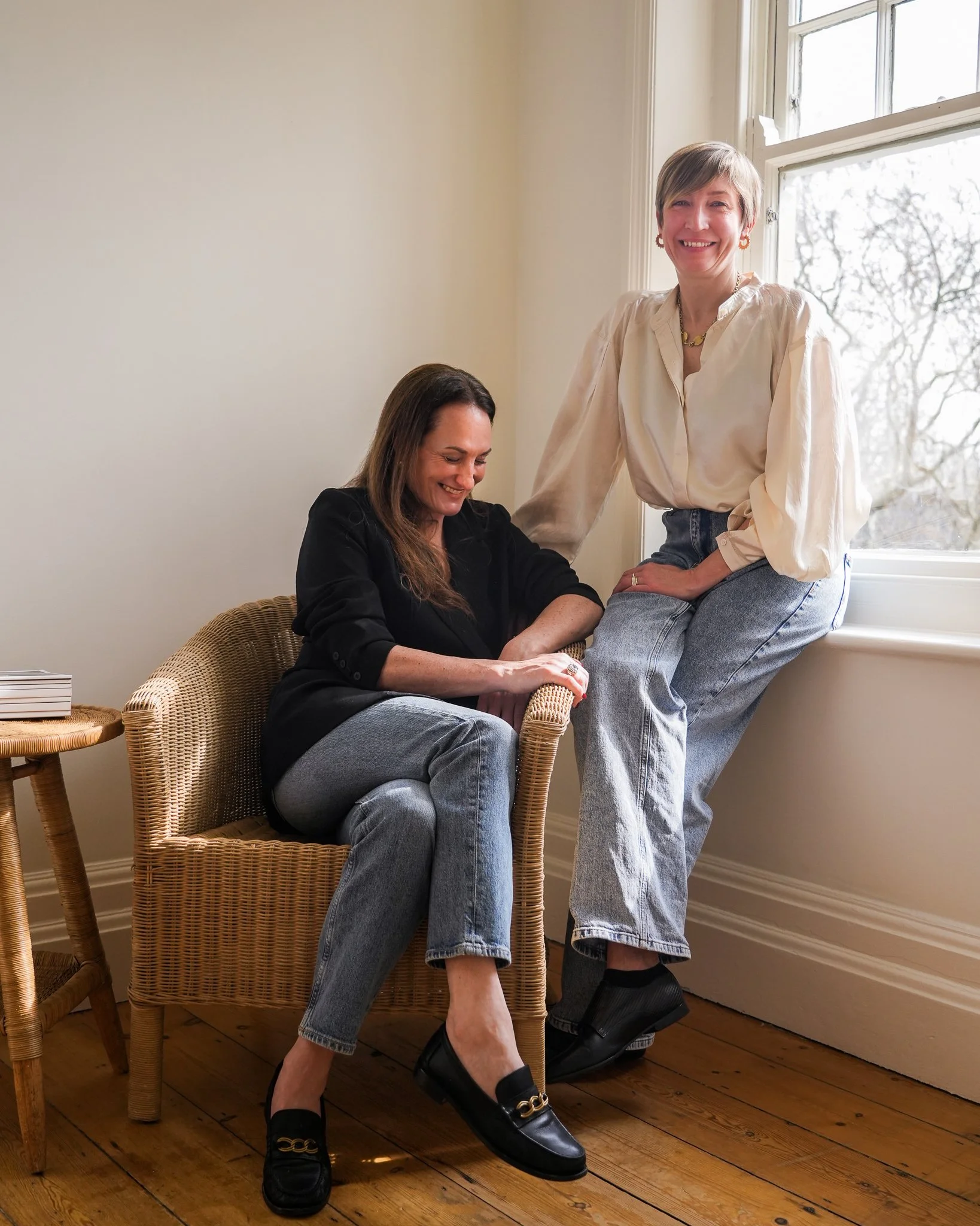 Two women smiling and laughing in a cozy room, one sitting on a wicker chair near a window, the other standing with her foot on the chair, both dressed in casual clothes.