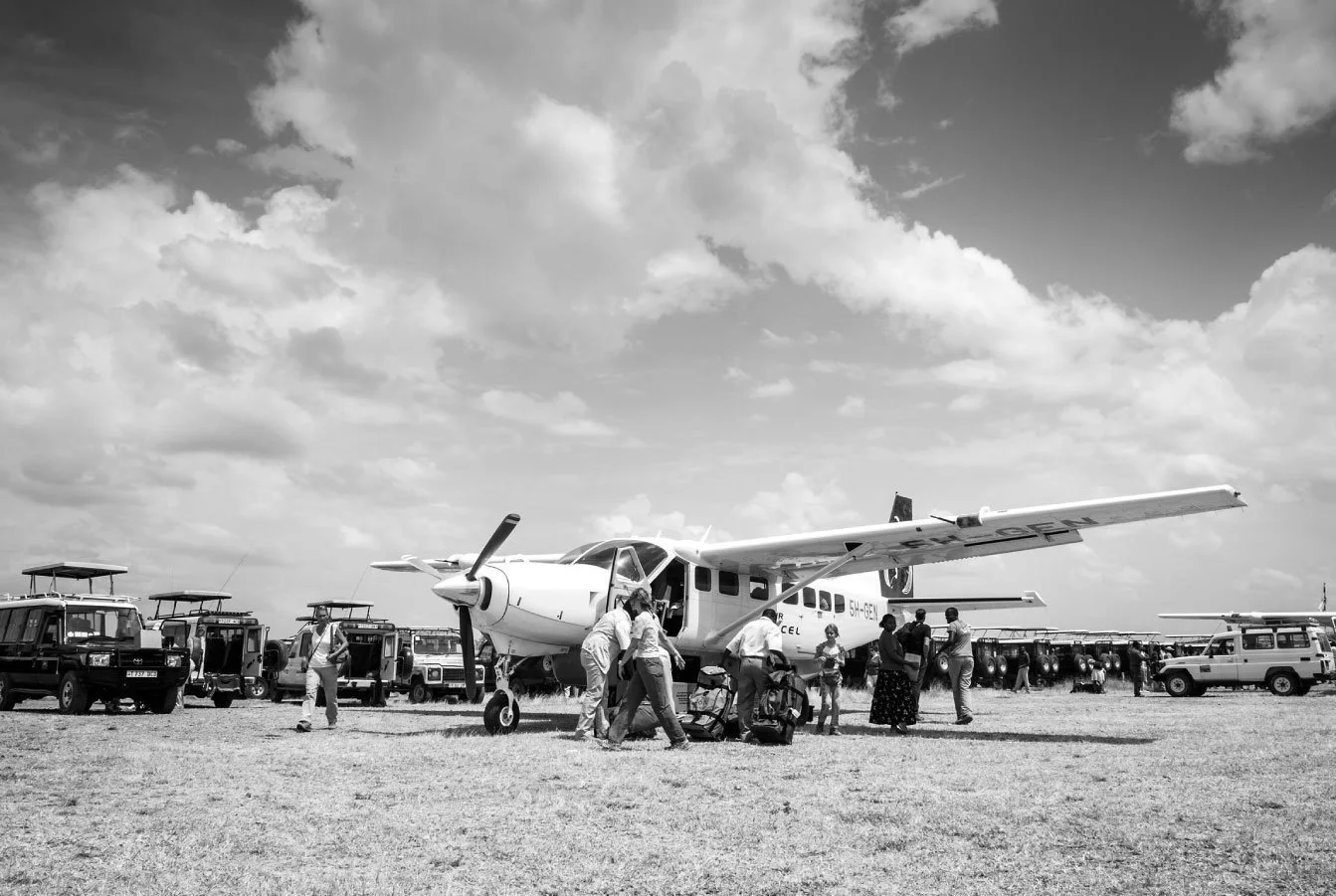A small aircraft on a grassy airfield with people loading luggage, surrounded by vehicles and parked planes, under partly cloudy skies.