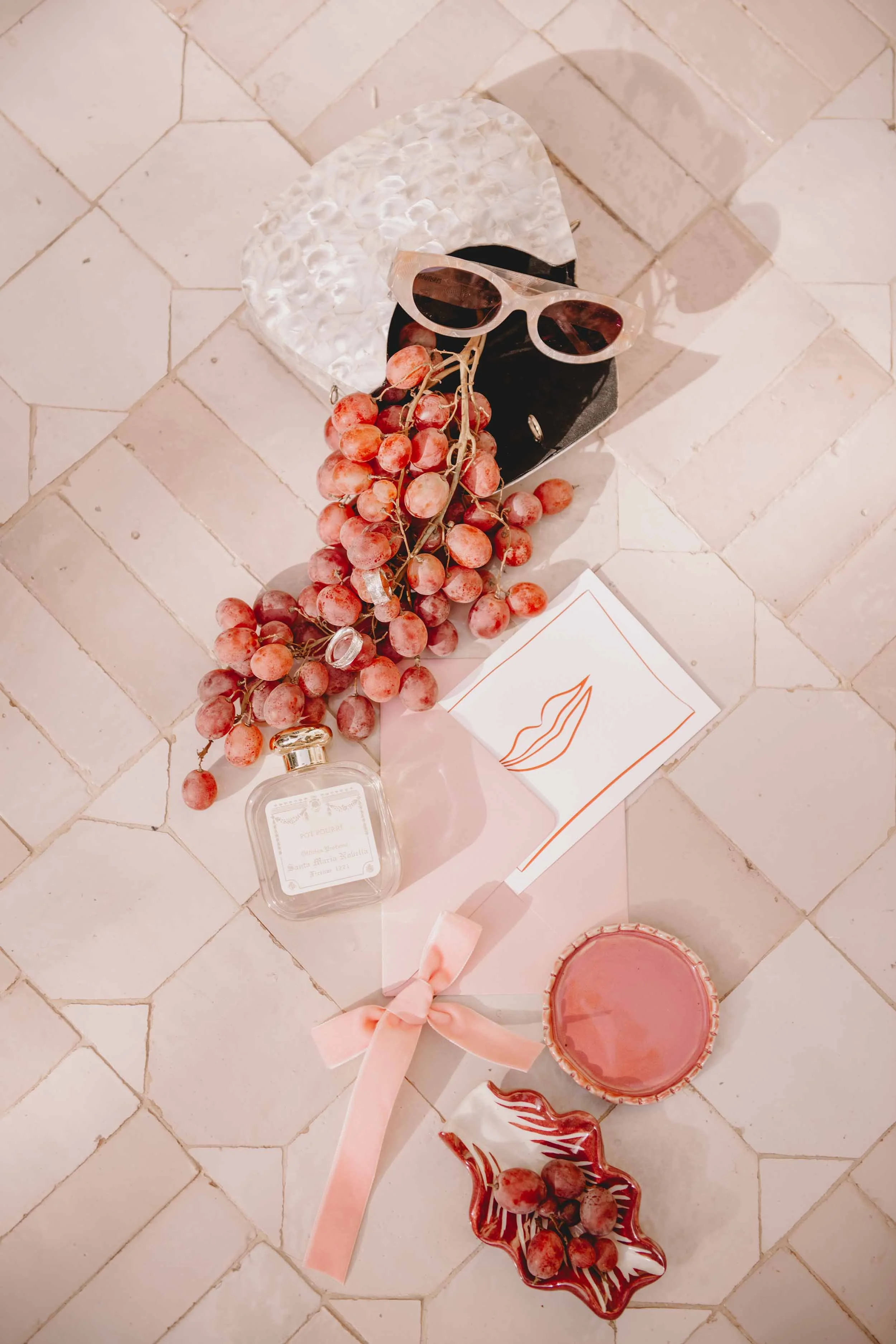 A flat lay arrangement on a tiled floor includes a pair of white sunglasses, a bunch of red grapes, a small jar of perfume, a pink ribboned paper cone with a lips illustration, a pink candle in a glass jar, and a small decorative dish with grapes.