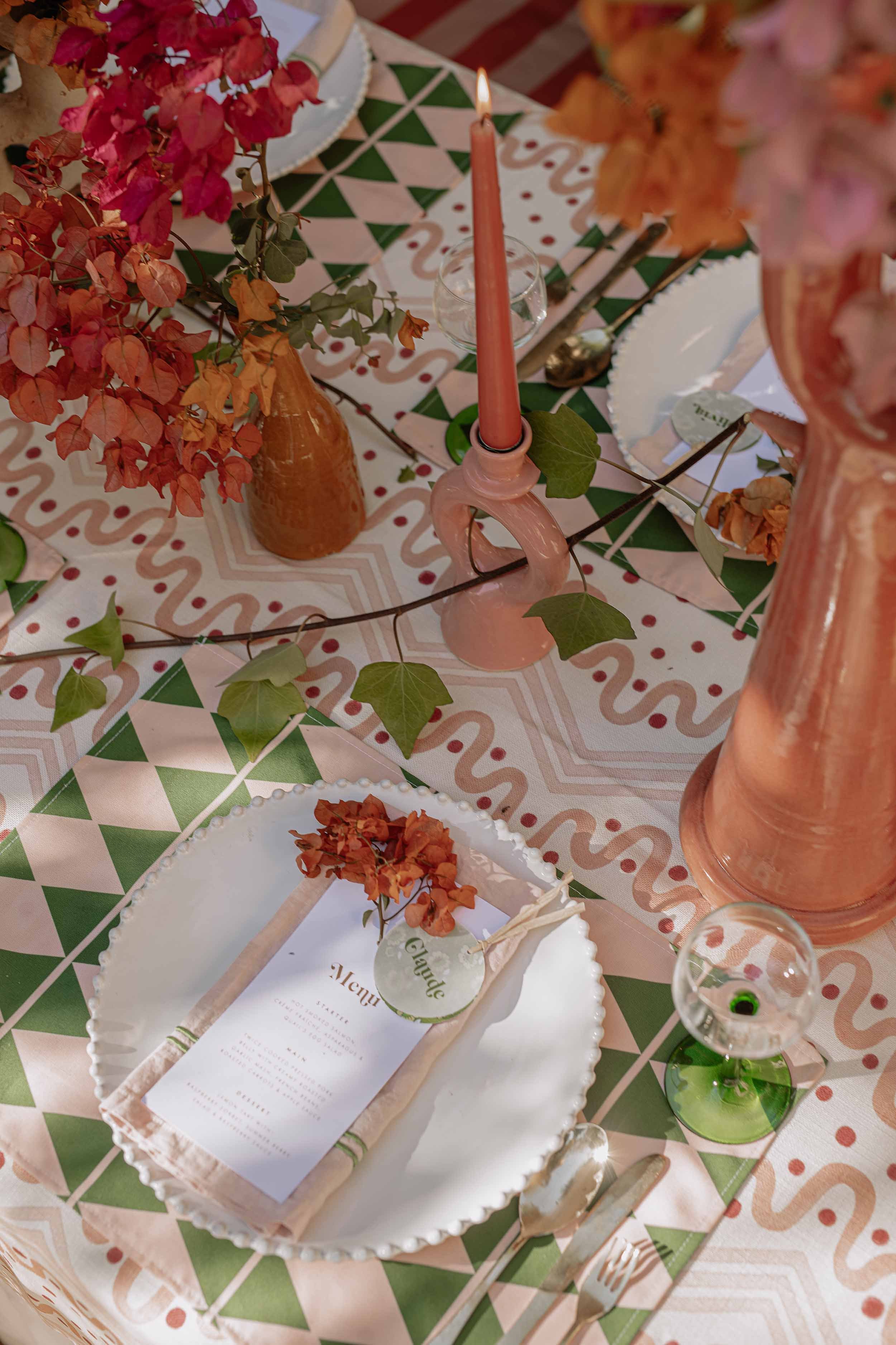 Decorated dining table with fall-themed centerpieces, pink and copper vases, pink candle, leafy garland, and a place setting with a menu, napkin, and glassware.