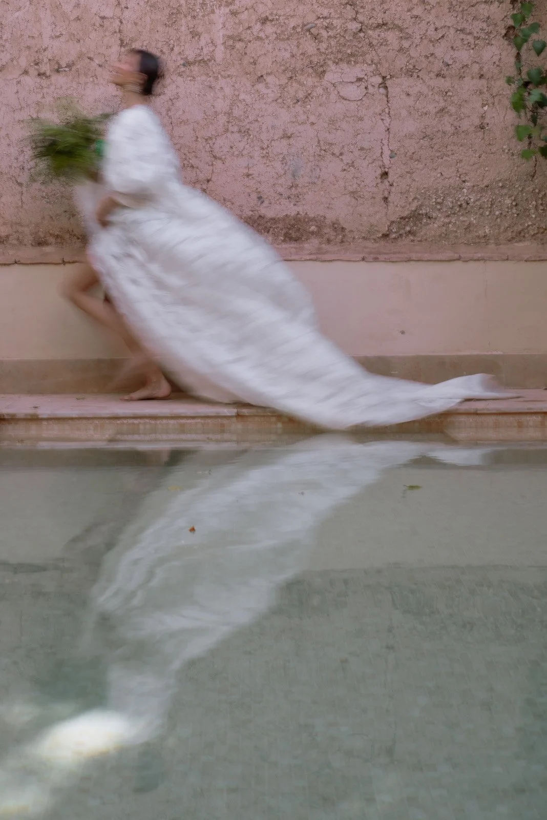 Bride in white dress with blurred motion, holding green plants, stepping along pool ledge with their reflection visible in the water.