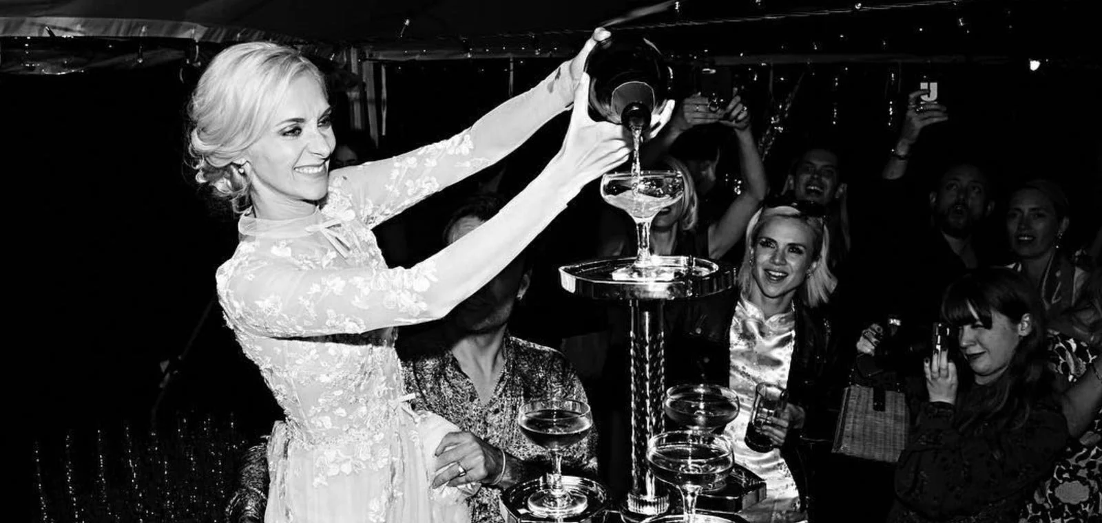 A woman in a white lace dress pours champagne into a tall glass at a celebration, surrounded by smiling guests in a festive setting.
