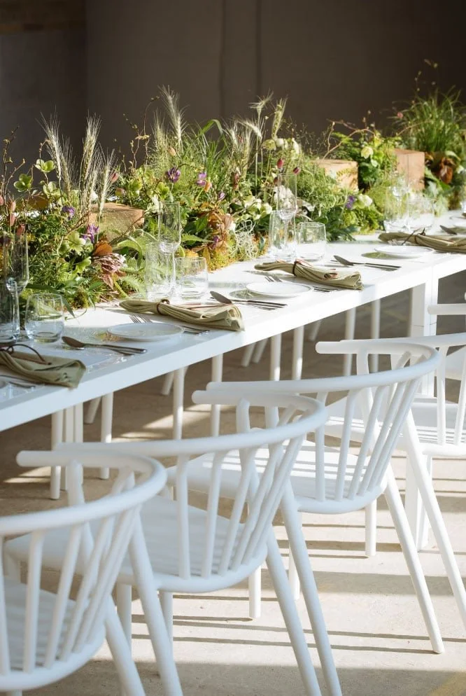 Long dining table with white chairs, decorated with green floral arrangements and table settings including napkins and glassware, in a well-lit room.